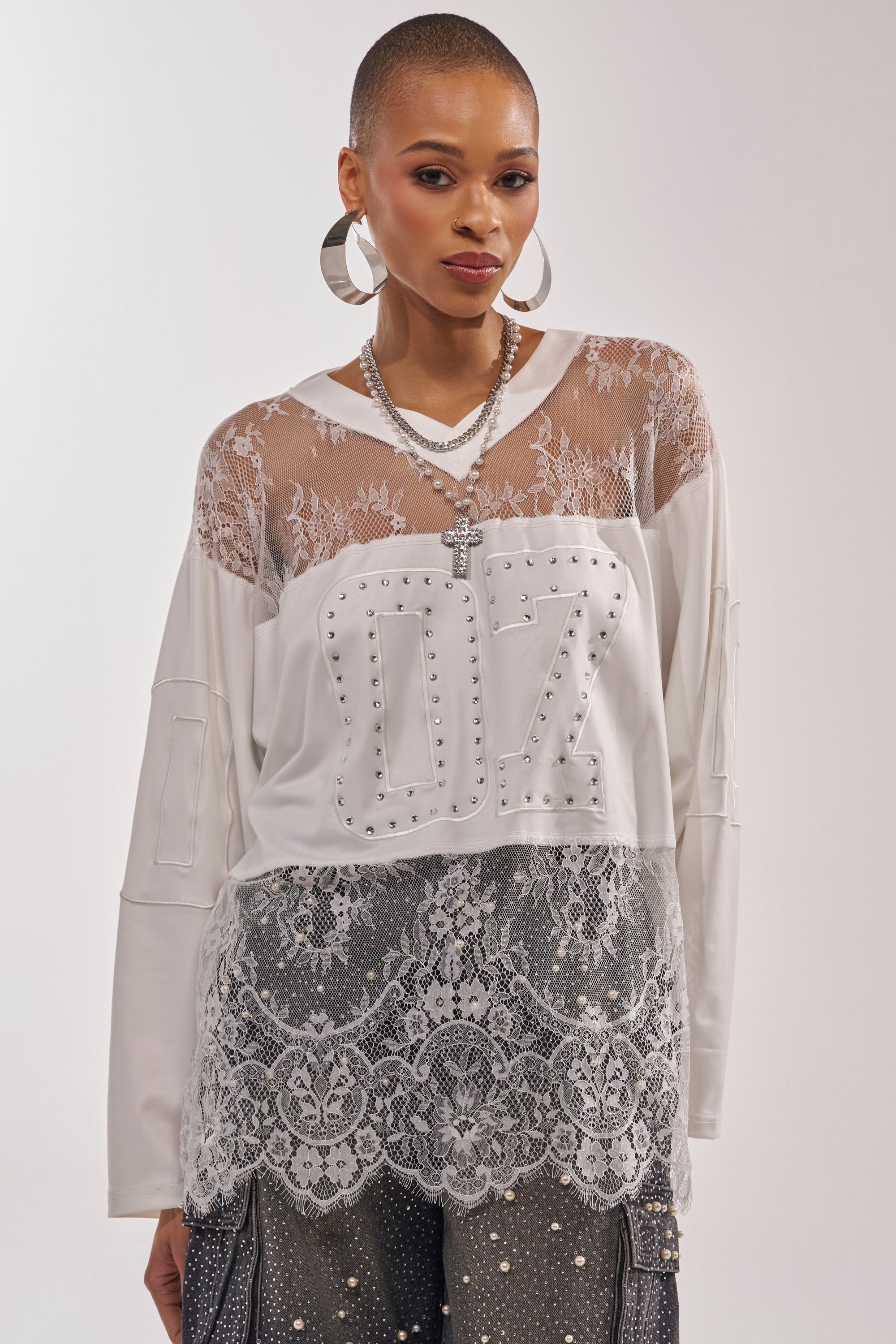 A woman with short hair wears hoop earrings, layered necklaces, and the WHO IS GETTING LUCKY TONIGHT JERSEY—a white long sleeve top with lace details—over a sheer lace shirt, paired with embellished pants. She stands against a plain background. 0