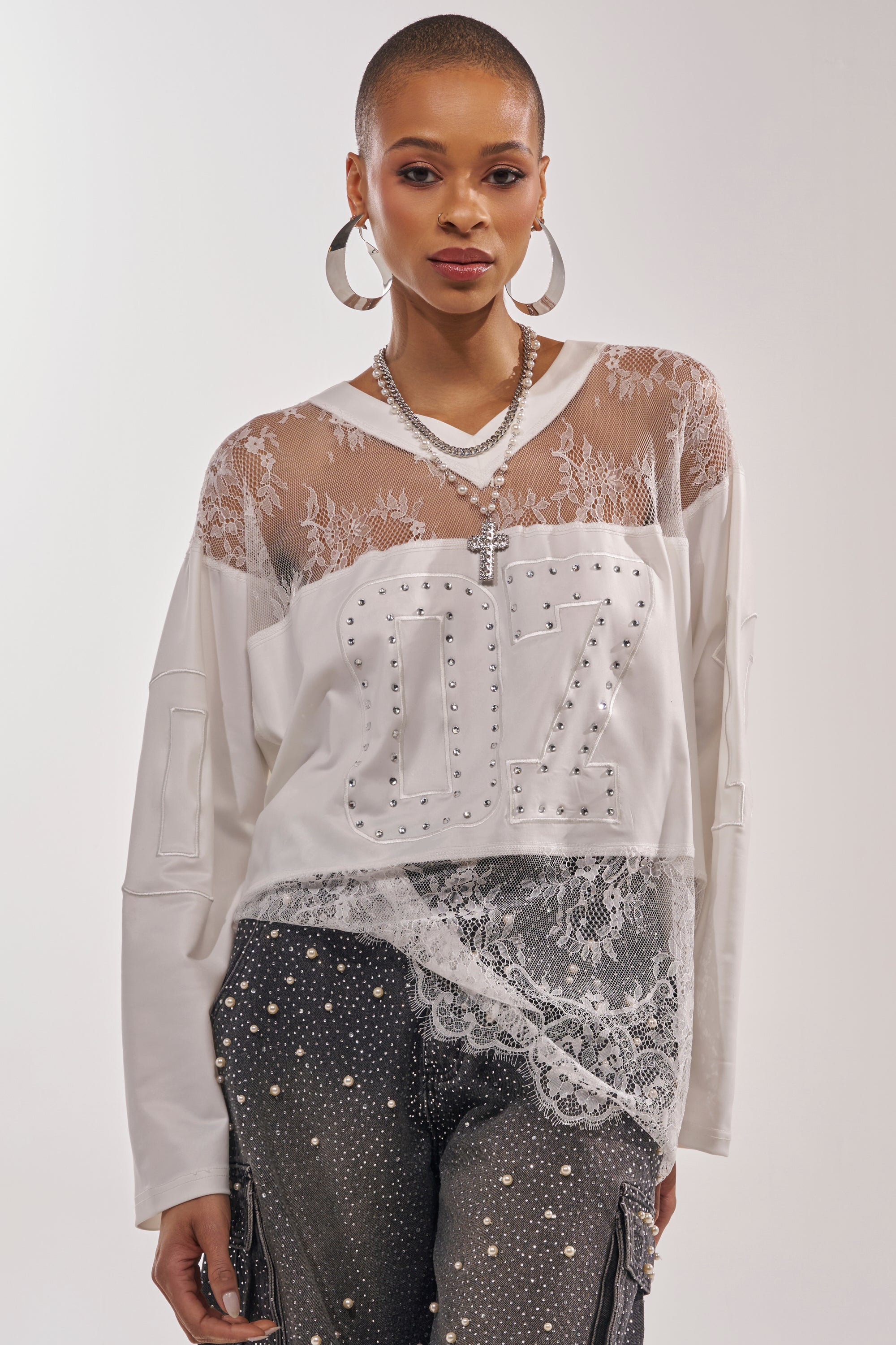 A stylish woman with a shaved head wears large hoops, layered necklaces, and the WHO IS GETTING LUCKY TONIGHT JERSEY—a white long-sleeve with lace and studded "07"—paired with high-waisted black pants featuring lace and studs. 1