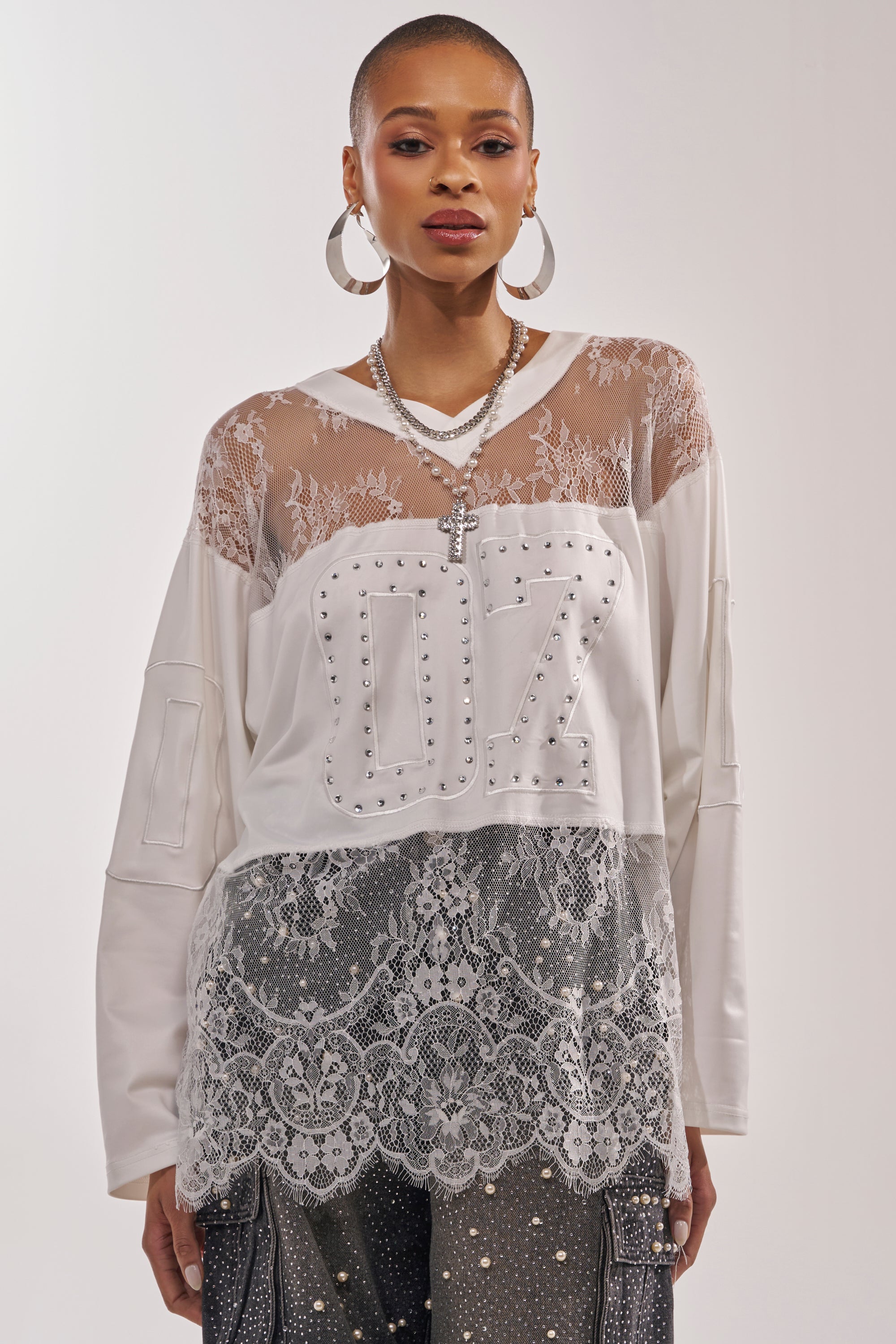 A woman wears the WHO IS GETTING LUCKY TONIGHT JERSEY, a white long sleeve lace top with sheer details and studded "07," paired with lace pants, hoop earrings, and layered necklaces as she poses against a plain background. 2