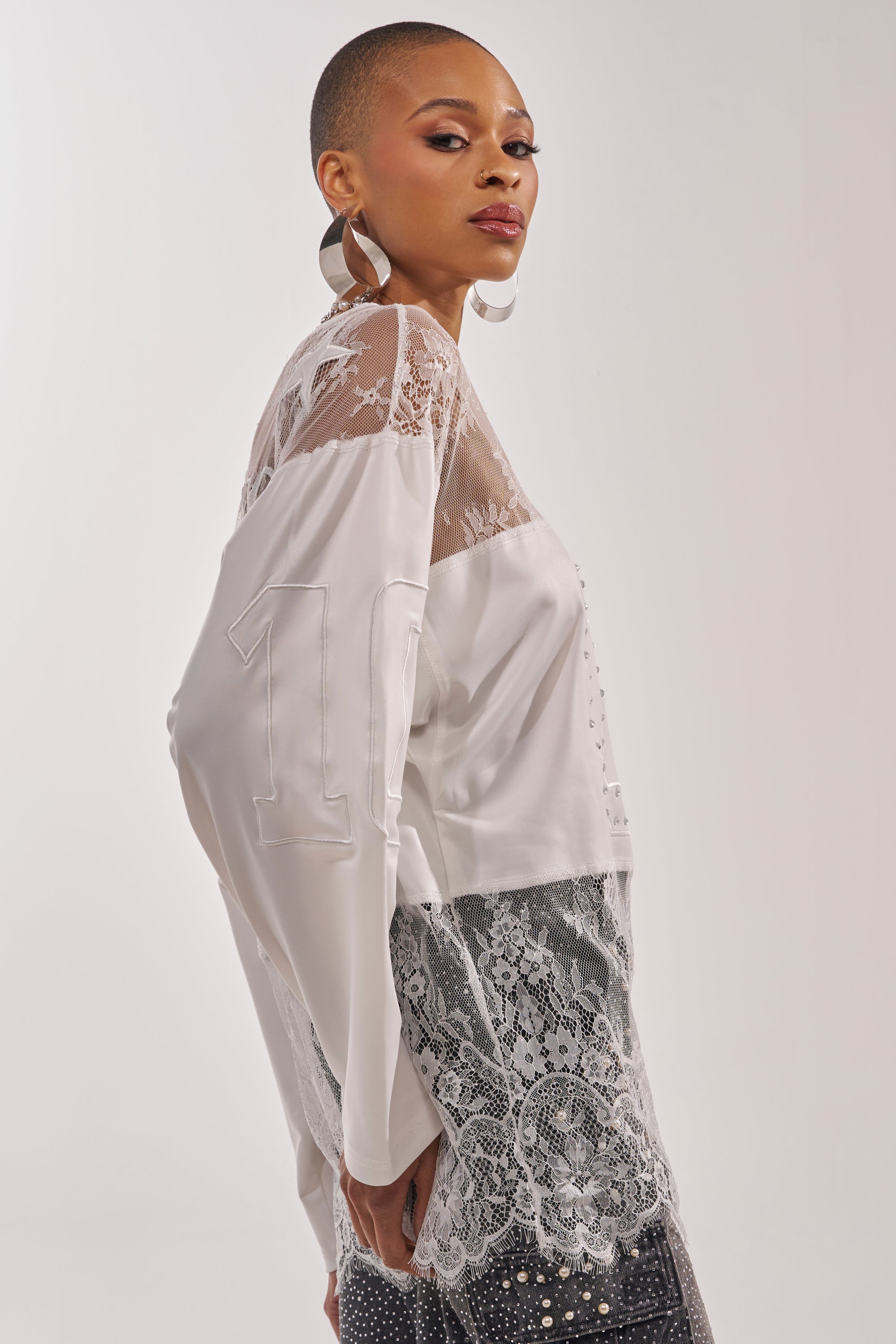 A person with short hair and large hoop earrings poses confidently in the WHO IS GETTING LUCKY TONIGHT JERSEY, a long sleeve white top featuring sheer lace panels and the number 10 on the sleeve, set against a plain light background. 3