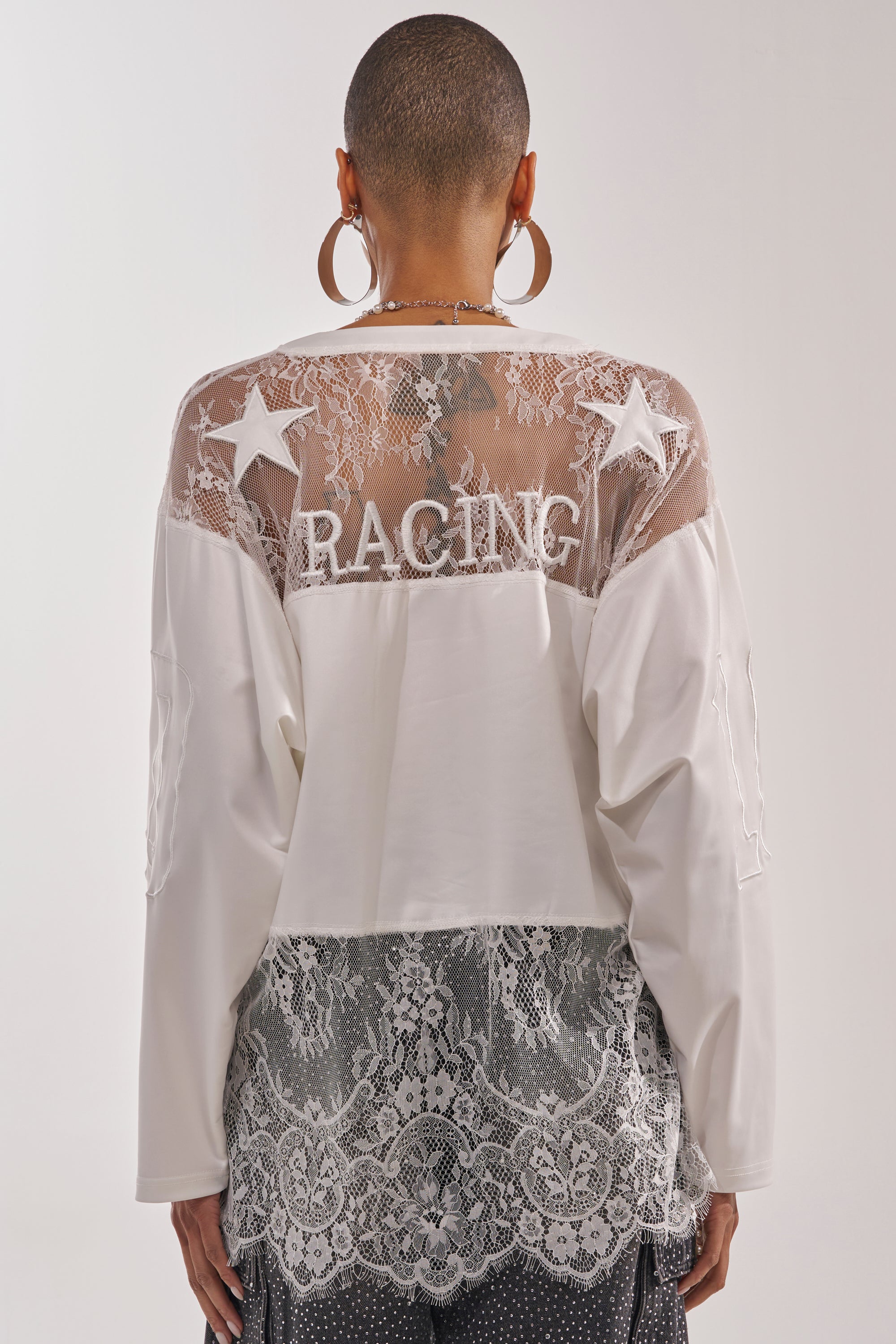 A person with short hair and hoop earrings is seen from behind, wearing the WHO IS GETTING LUCKY TONIGHT JERSEY, featuring lace fabric, sheer panels, star details, and bold “RACING” embroidery across the back. 4