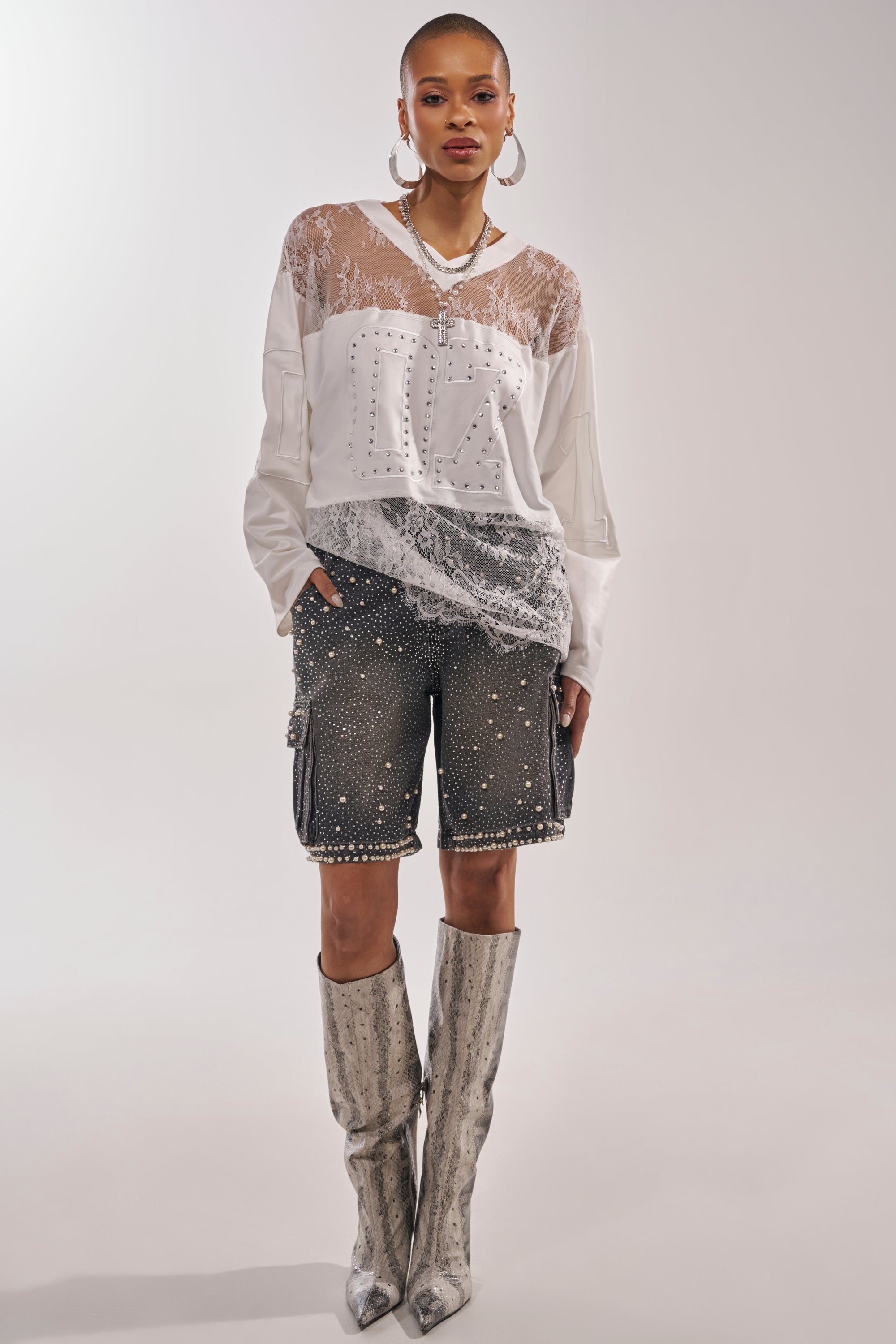 A stylish woman poses in the WHO IS GETTING LUCKY TONIGHT JERSEY, paired with black studded shorts and silver knee-high boots. She completes the look with large hoop earrings and layered necklaces against a light background. 5