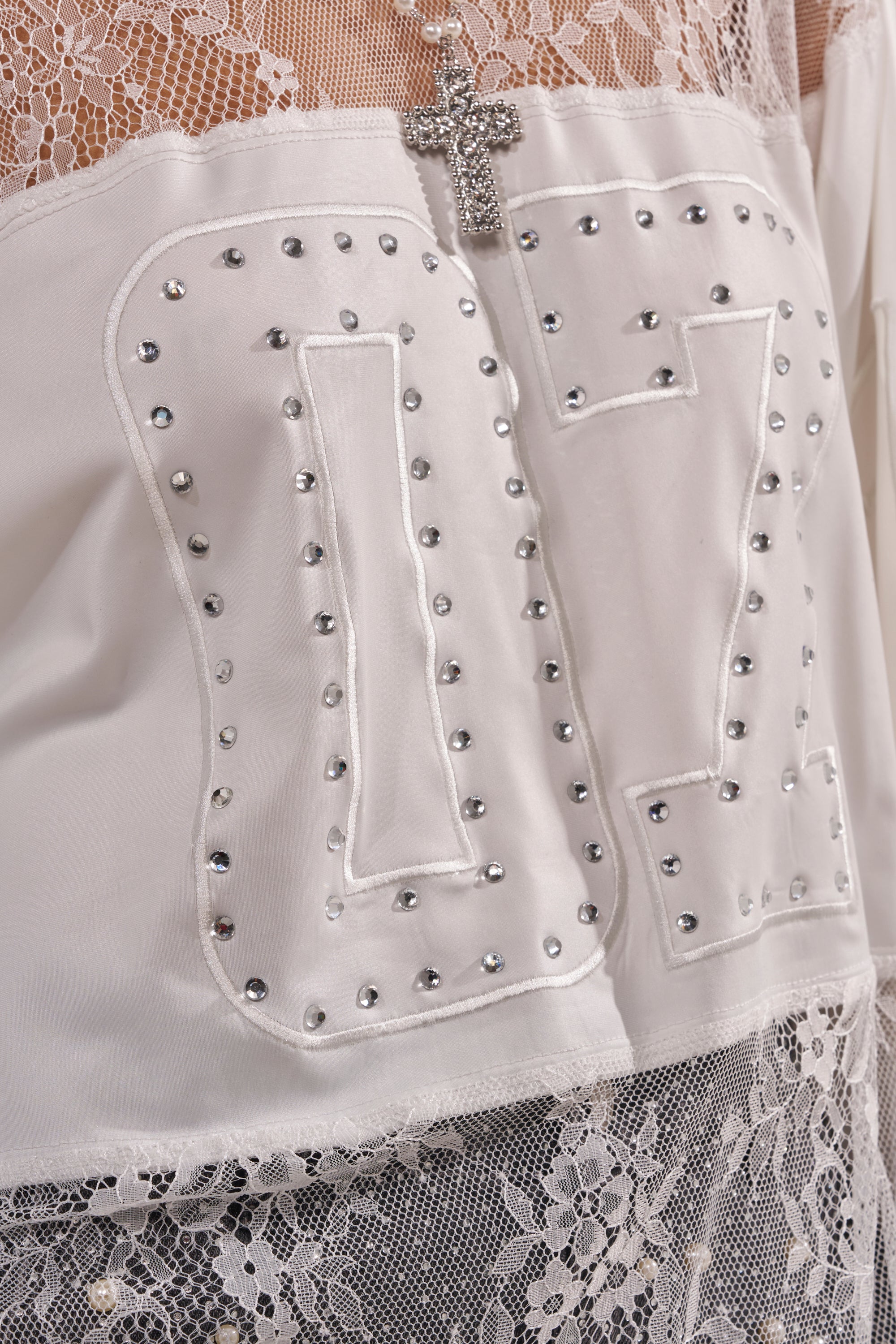 Close-up of the WHO IS GETTING LUCKY TONIGHT JERSEY: a white lace jersey top featuring a sparkly cross pendant, stud-outlined "17" on satin, and mesh and lace details adding texture at the neckline and hem. 6