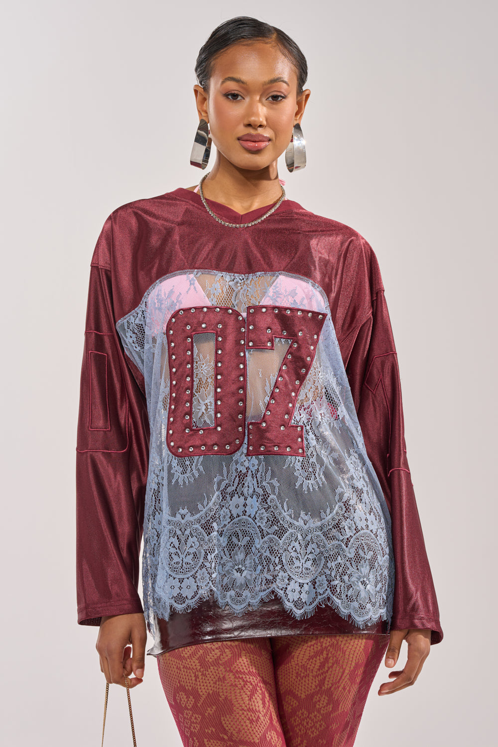 A woman poses confidently in the WINNER JERSEY—a sporty-chic burgundy lace top with bold "07" studs and a sheer blue panel—paired with red patterned pants and large hoop earrings. 0