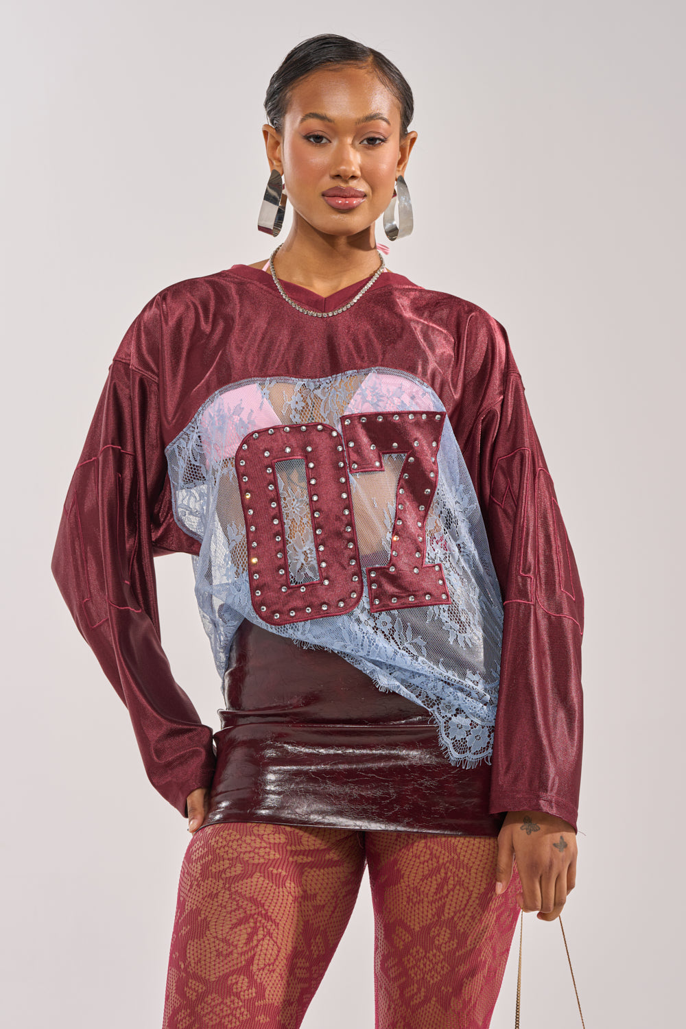 A woman stands confidently against a plain background, wearing the WINNER JERSEY—a sporty-chic burgundy lace top with "07" on the front—paired with a shiny skirt, patterned tights, hoop earrings, and styled hair. 1