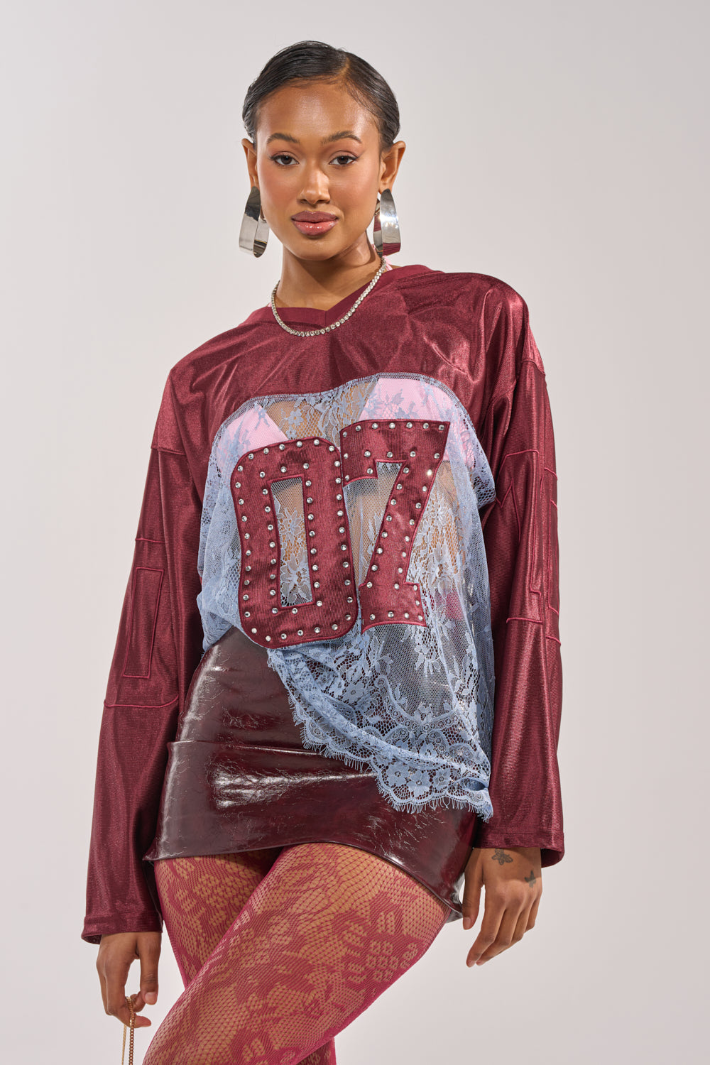A woman poses confidently in the sporty-chic WINNER JERSEY, featuring bold "OZ" letters, paired with hoop earrings, a shiny maroon skirt, and pink patterned tights against a plain background. 2