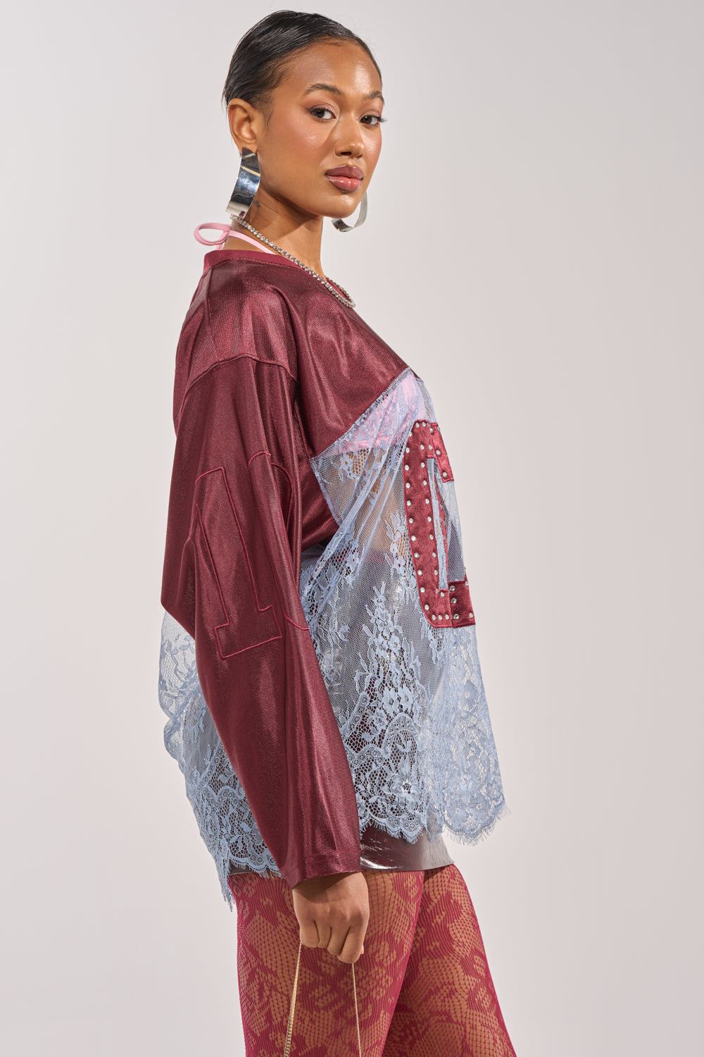 A woman models the WINNER JERSEY, a maroon and sheer blue lace top with the number 10 on the sleeve, paired with large hoop earrings and red patterned tights for a sporty-chic look against a plain background. 3