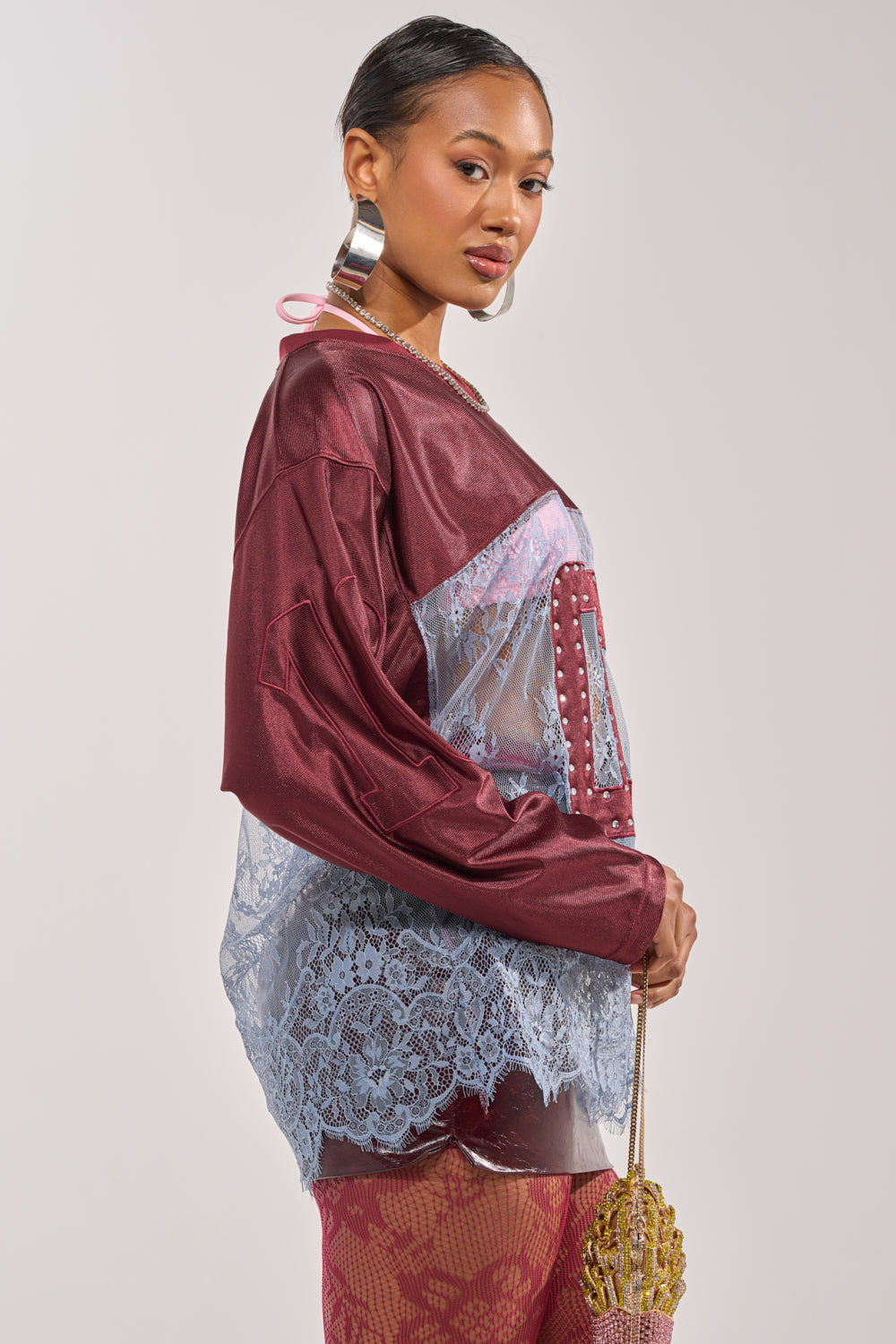 A woman poses in the WINNER JERSEY, a sheer blue lace blouse layered under a maroon satin top, styled with patterned maroon tights, large hoop earrings, and a gold fan-shaped clutch. 4