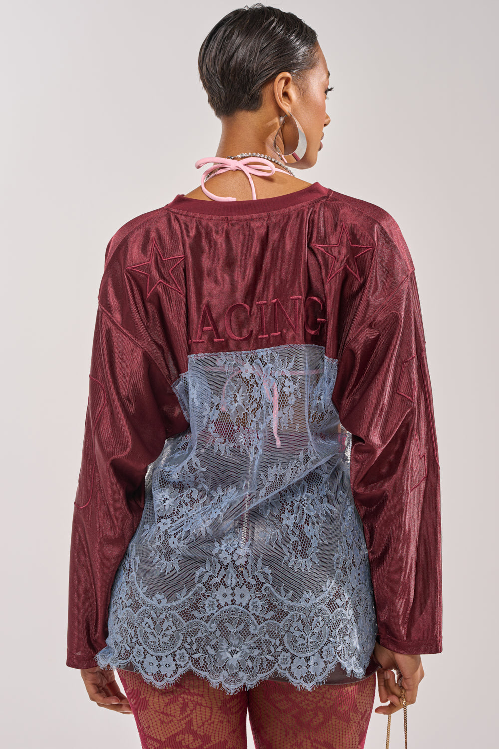 A woman faces away from the camera wearing the WINNER JERSEY: a maroon lace top with star details and "RACING" text, featuring sheer blue lace layers and a pink halter strap for a sporty-chic, mixed media look. 5