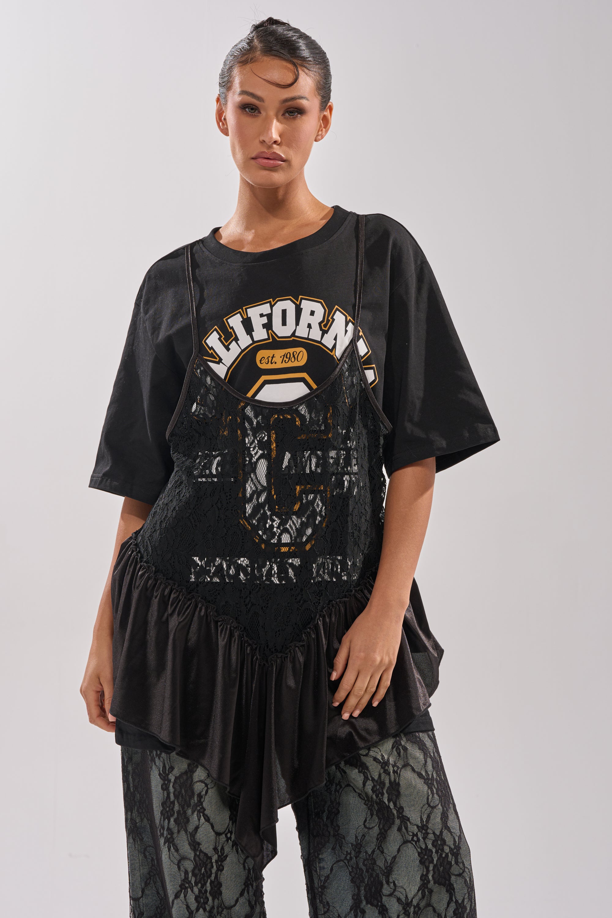 A woman faces forward in the BEVERLY HILLS DIVA LACE OVERLAY TEE, layered over a black lace dress and matching lace pants for a sporty-chic streetwear look. She poses with slicked-back hair against a plain light background.