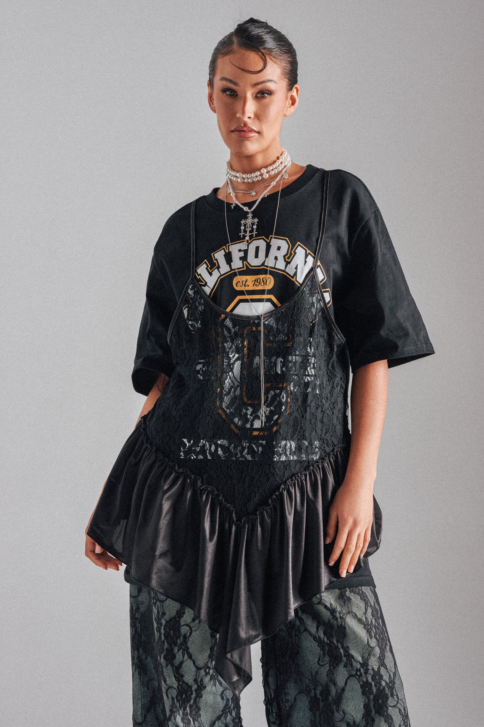 A woman confidently poses against a gray background wearing the BEVERLY HILLS DIVA LACE OVERLAY TEE, layered with a black lace and satin dress, dark lace pants, and multiple necklaces.