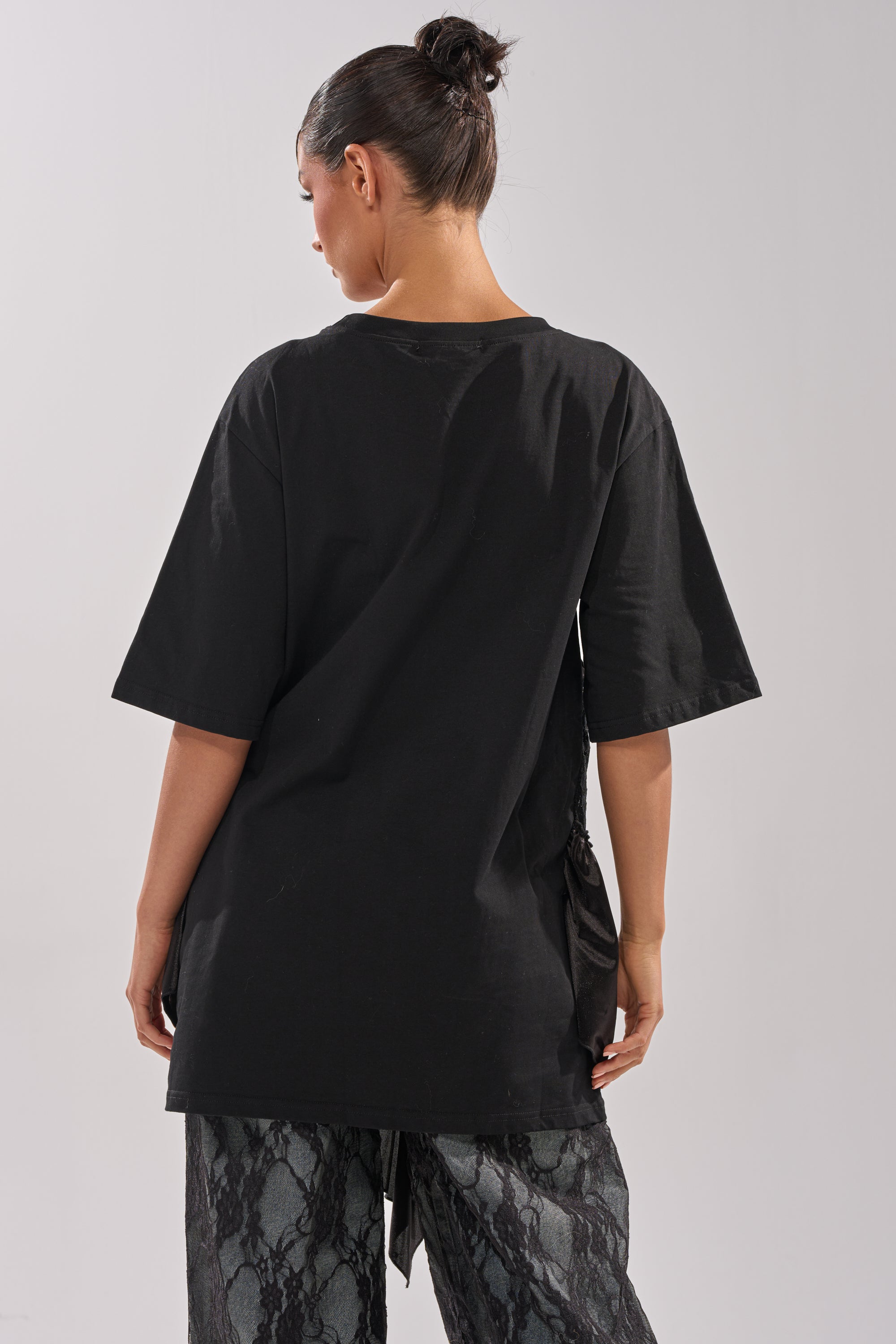 A person with dark hair in a bun stands with their back to the camera, wearing the BEVERLY HILLS DIVA LACE OVERLAY TEE and dark patterned pants against a plain light background.