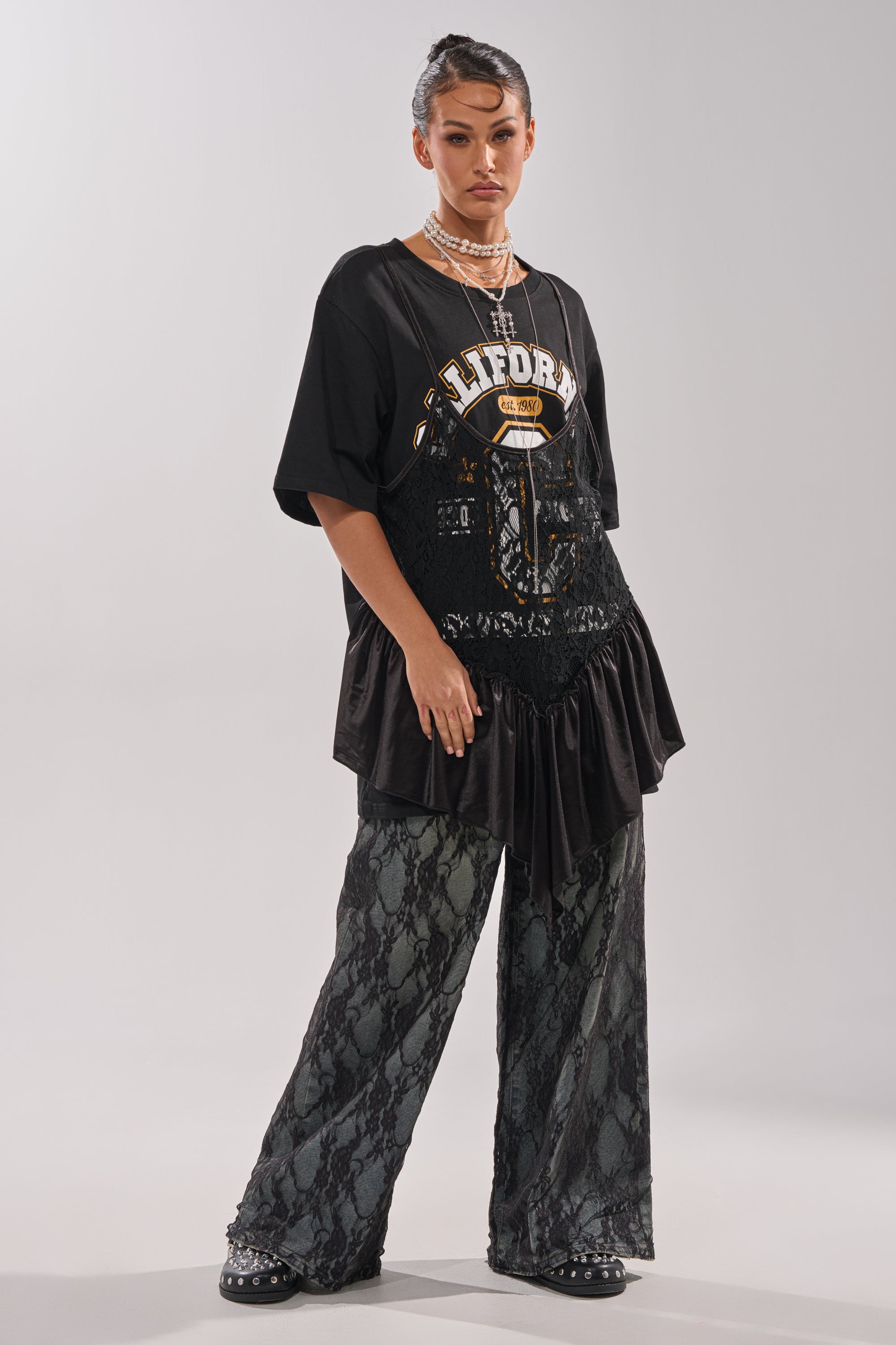 A person faces forward against a plain background, wearing the BEVERLY HILLS DIVA LACE OVERLAY TEE styled over a black lace dress and wide lace pants, with chunky black shoes, layered necklaces, and a slicked-back hairstyle.