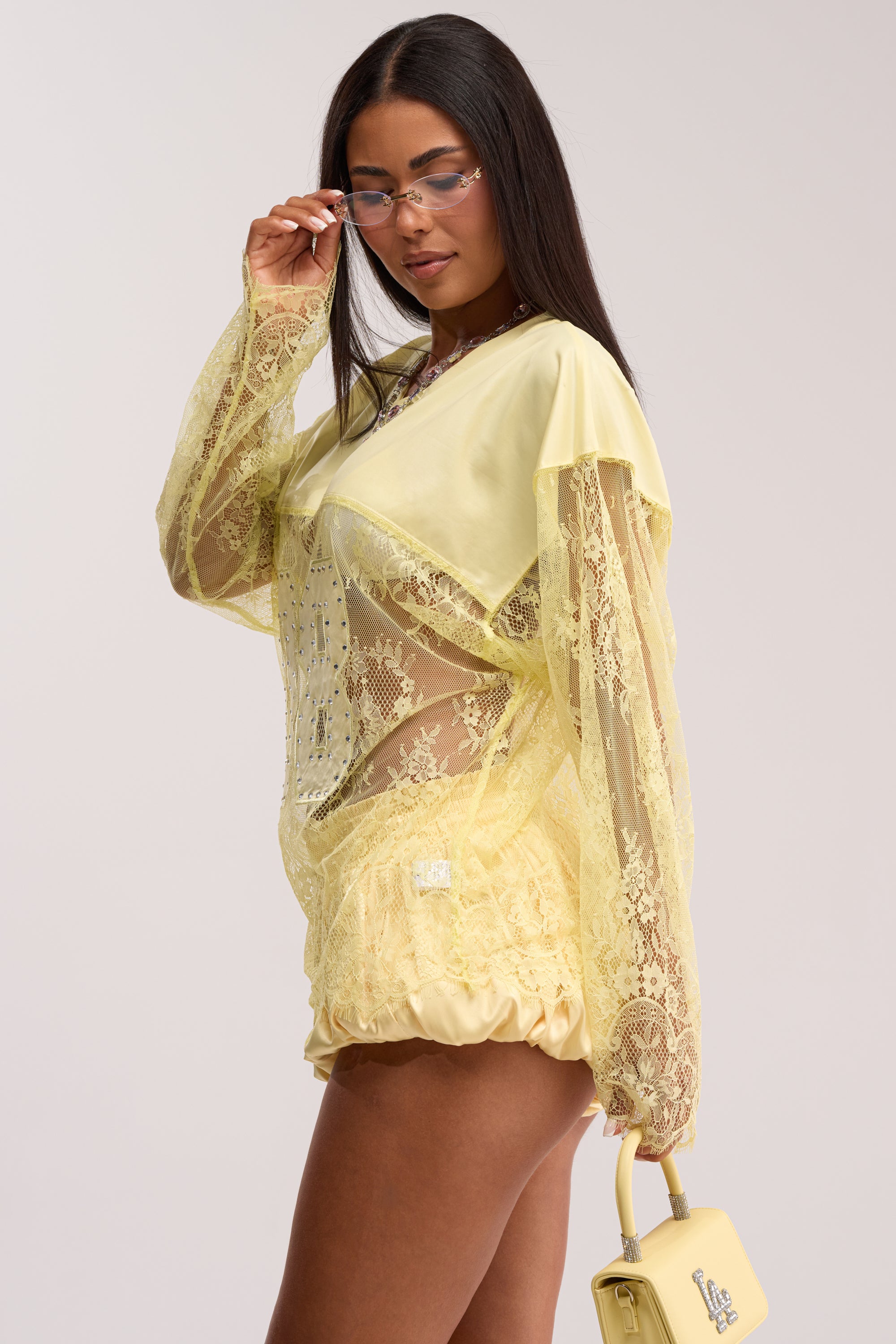 A woman with long dark hair wears the GIRLS ARE PLAYERS TOO LACE JERSEY TOP IN YELLOW and matching shorts, holding a small yellow handbag. She adjusts her clear glasses while standing against a plain light background.