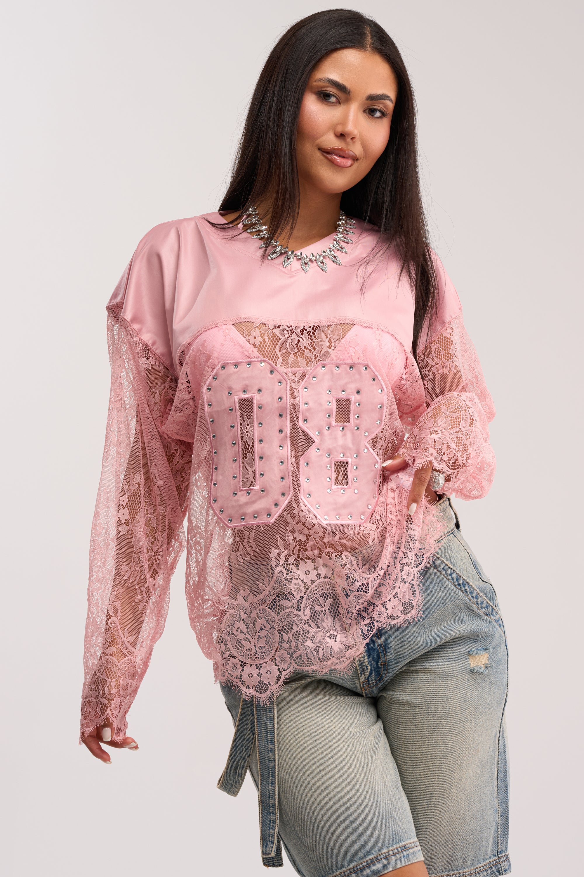 A woman with long dark hair confidently poses in the GIRLS ARE PLAYERS TOO LACE JERSEY TOP—a pink jersey with "08" and rhinestone patches—over a pink shirt, paired with light denim shorts, one hand on her hip.
