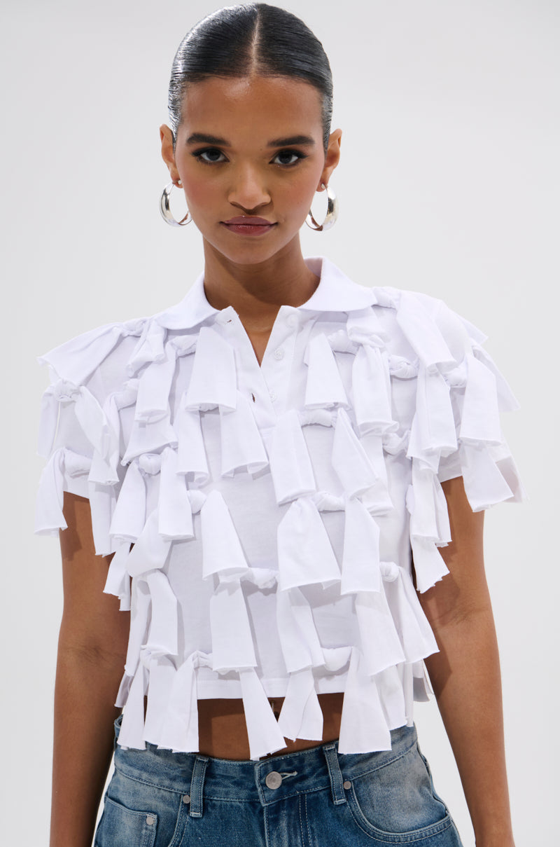 A woman wearing hoop earrings, blue jeans, and the TIED DOWN KNOT TOP—a white collared short-sleeve top with 3D tassel details—stands against a plain light background.