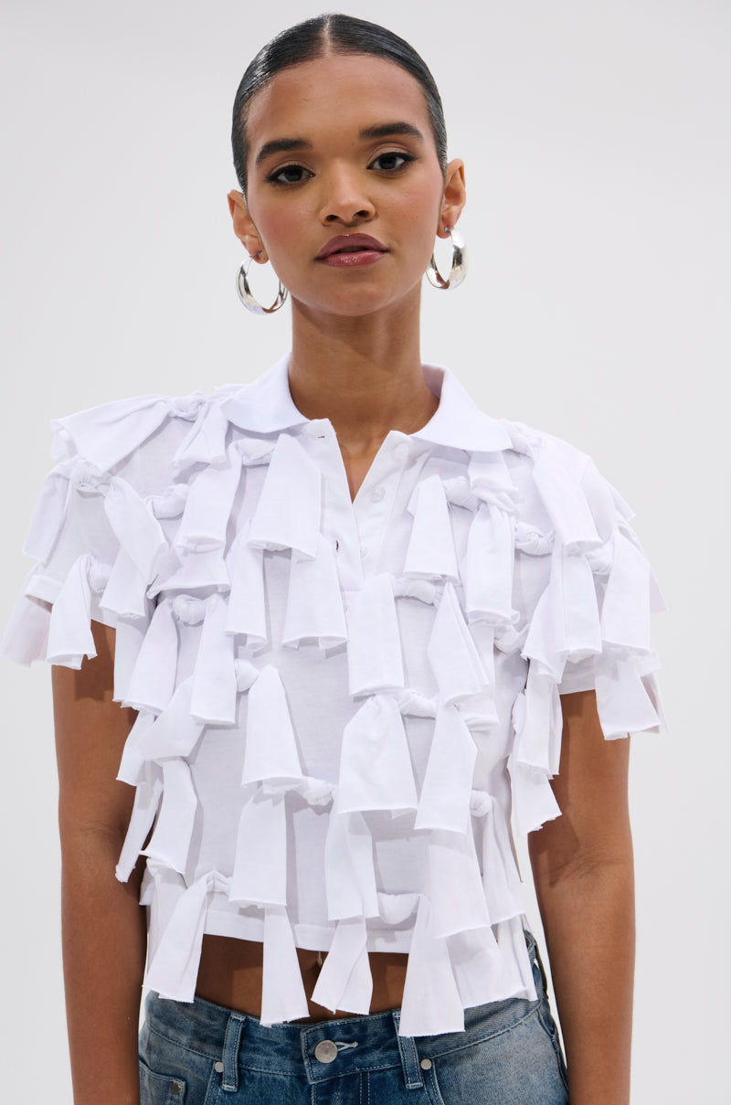 A woman models the TIED DOWN KNOT TOP—white, short-sleeve, with layered fabric strips and a relaxed cropped fit—paired with blue jeans, standing before a plain light background.