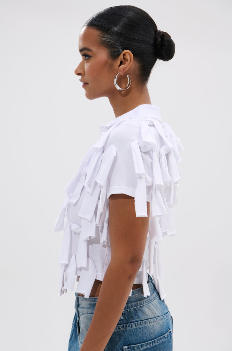A person with dark hair in a sleek bun models the TIED DOWN KNOT TOP, a white cropped top with layered fabric strips, paired with blue jeans and shown in profile against a plain background.