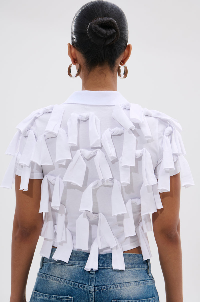 A person with hair in a neat bun and large hoop earrings wears the TIED DOWN KNOT TOP—a white cropped top with fabric strips at the back—paired with blue jeans, photographed from behind against a plain background.