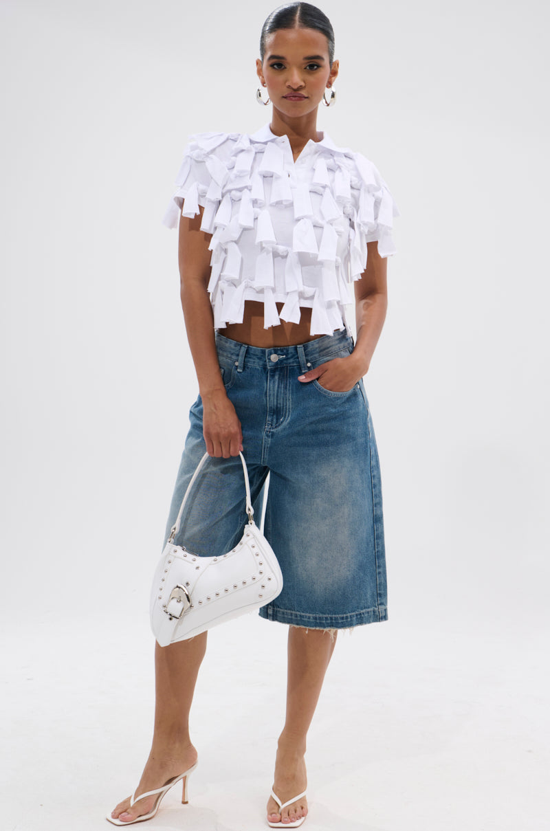 A woman stands confidently in the TIED DOWN KNOT TOP, styled with knee-length blue denim shorts, white heeled sandals, a white handbag, hoop earrings, and her hair tied back against a plain white background.