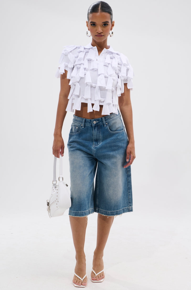A woman with pulled-back hair stands against a white background wearing the TIED DOWN KNOT TOP, statement earrings, knee-length denim shorts, white heeled sandals, and holding a white handbag, showcasing a chic cropped silhouette.