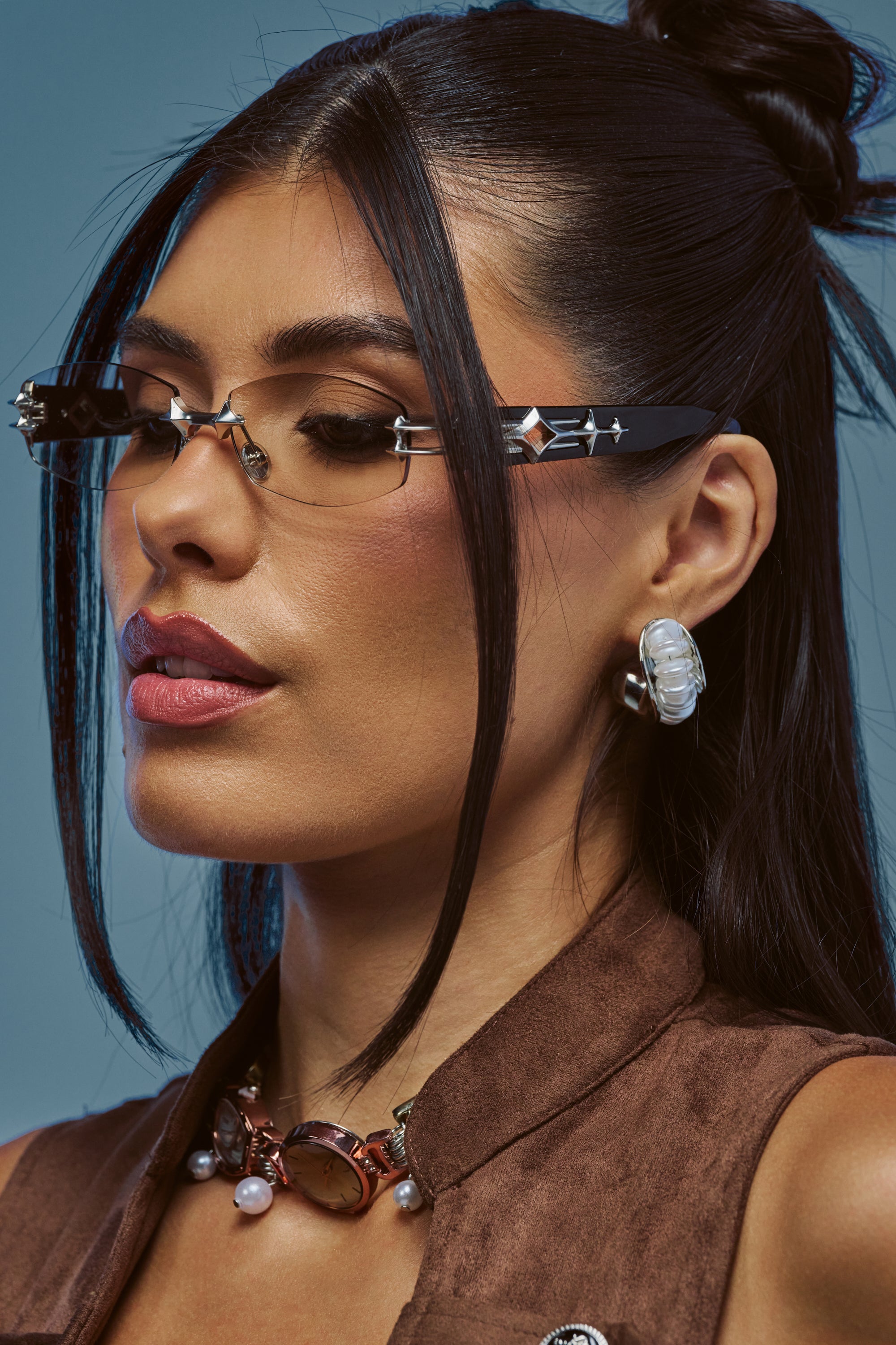 Woman with long dark hair and geometric glasses, wearing a brown suede top, the BY THE SHORE EARRING—wavy silver drop earrings—and layered necklaces adorned with pearlescent beads, looking down against a blue background.