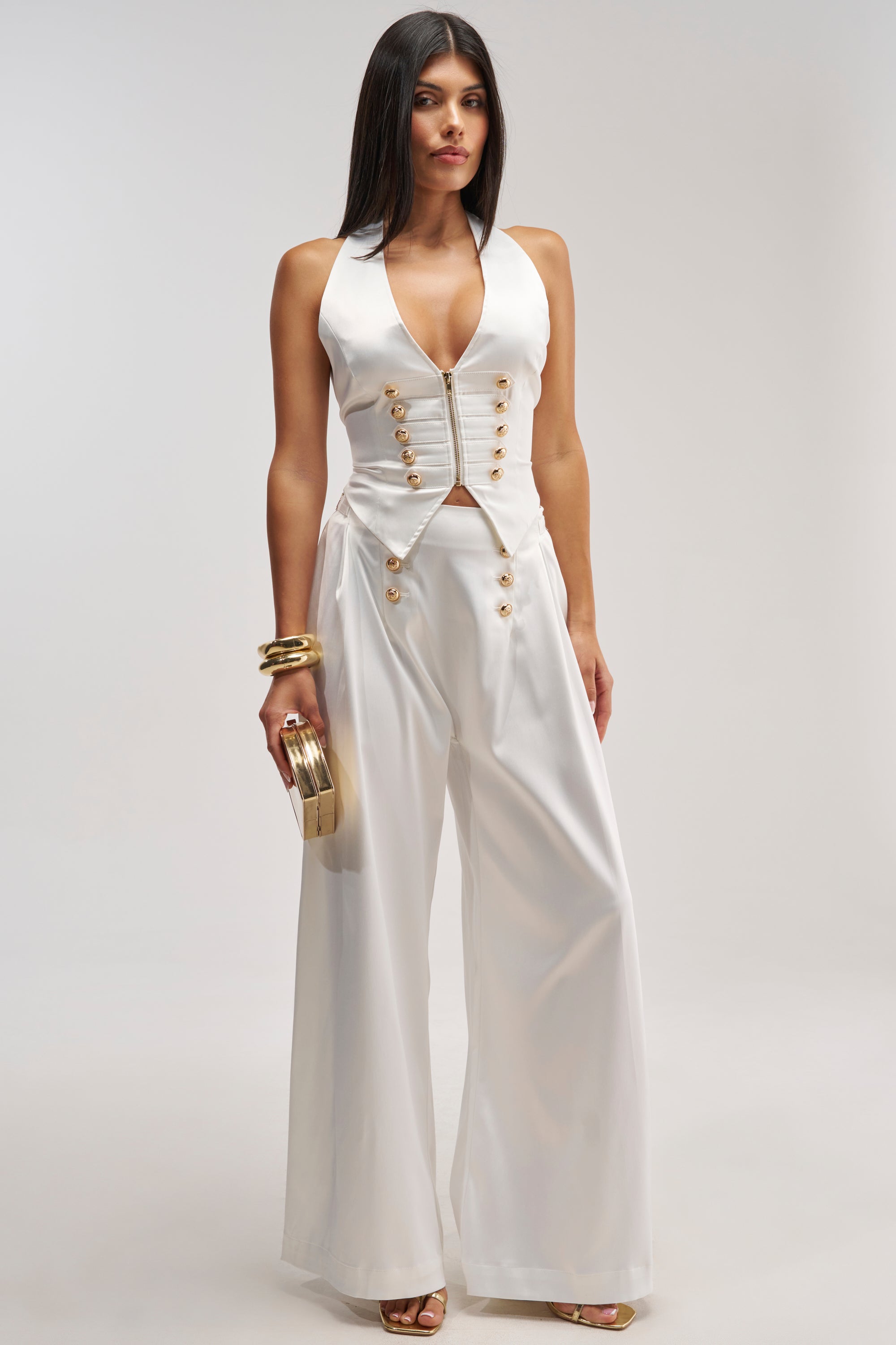 A woman stands against a plain light background wearing the WHITE PARTY EMBELLISHED RELAXED PANT, paired with a sleeveless white vest with gold buttons. She accessorizes with gold bracelets, heels, and a gold clutch.