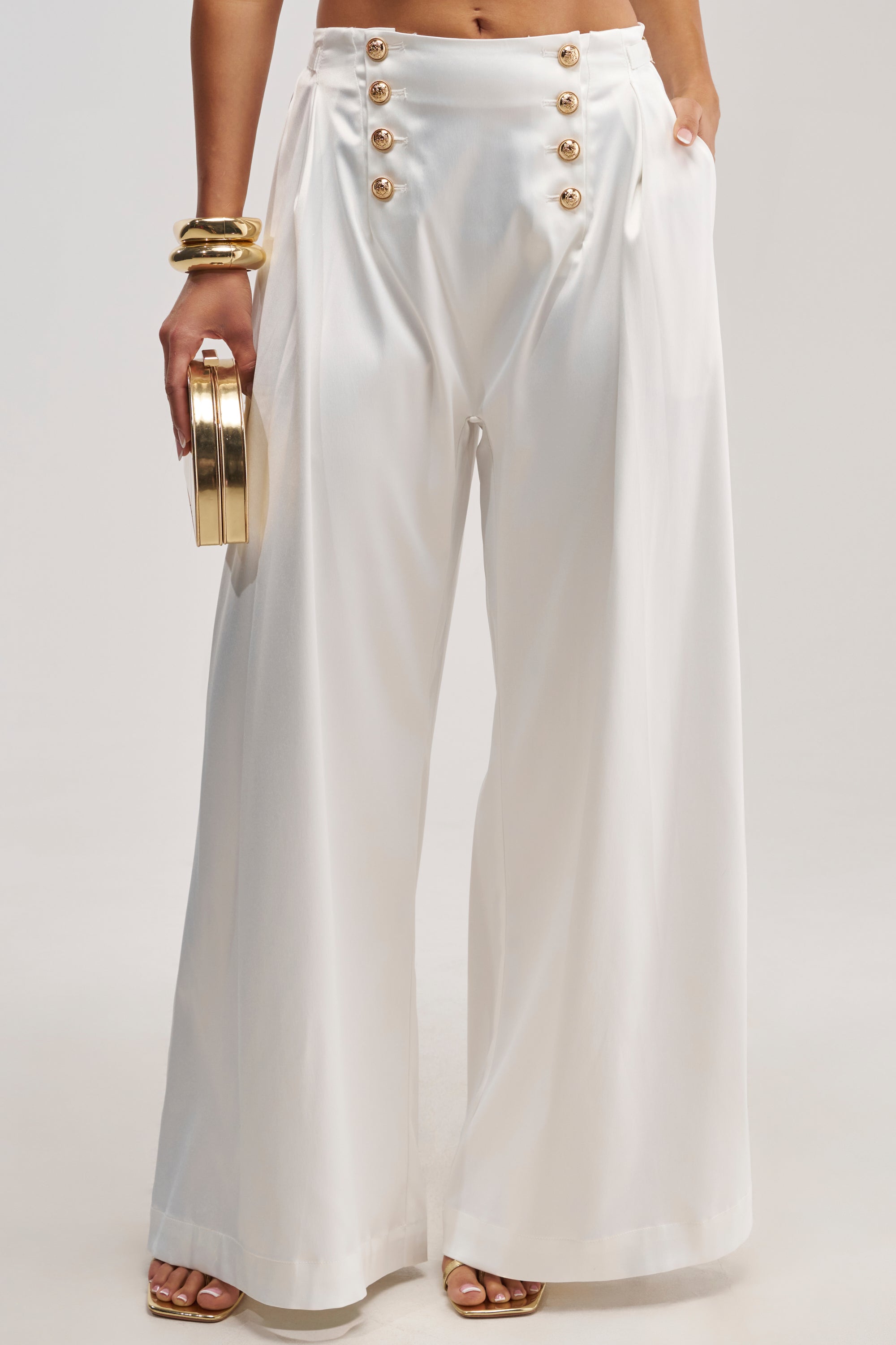 Dressed in the WHITE PARTY EMBELLISHED RELAXED PANT, this woman exudes elegance with its white satin wide leg design and striking gold hardware details. She completes her chic bridal look with a gold clutch, gold bracelets, and matching sandals.