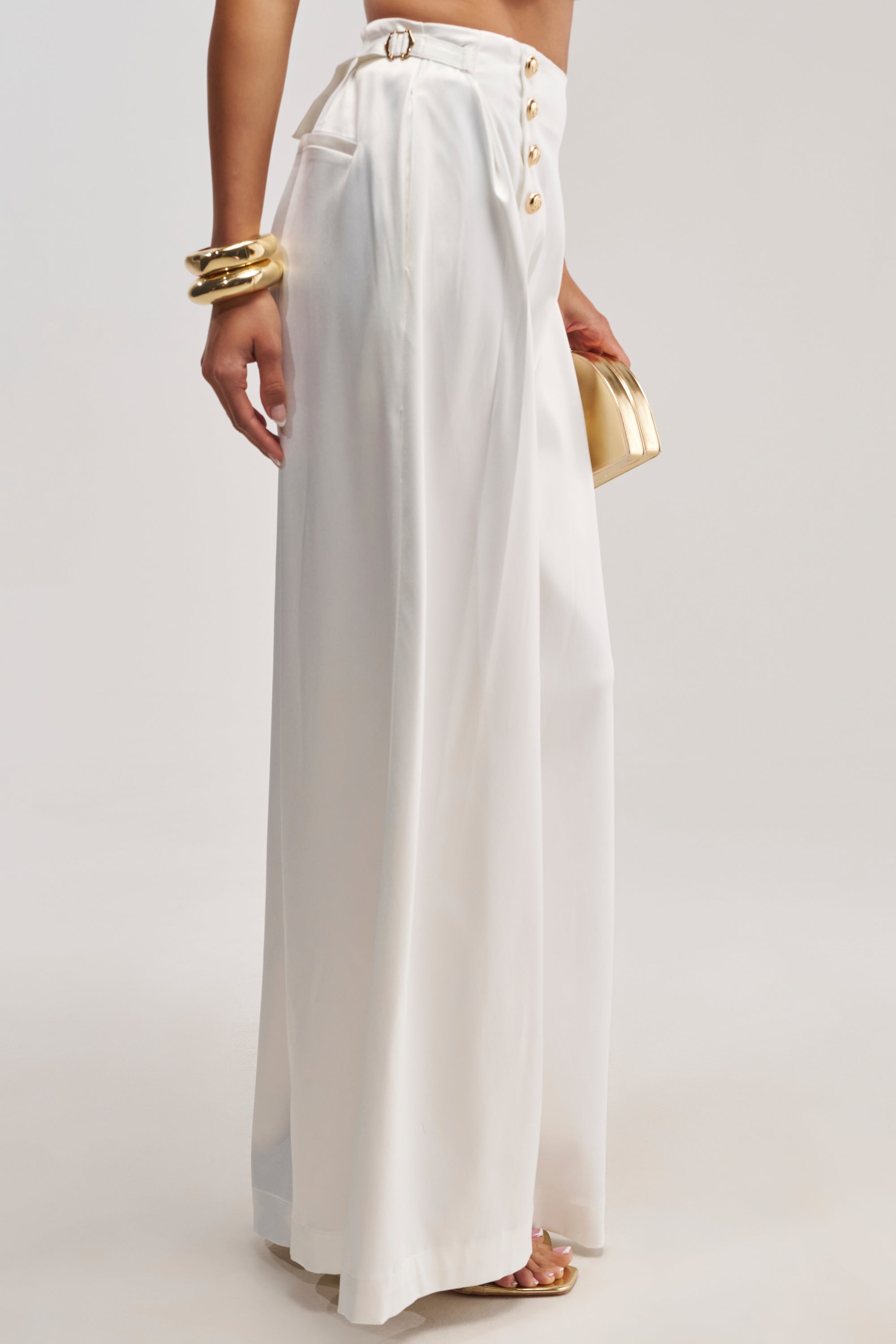 A person is shown from the waist down, wearing the WHITE PARTY EMBELLISHED RELAXED PANT—high-waisted, white satin wide leg pants with gold buttons—styled with a small gold clutch and chunky gold bracelets.