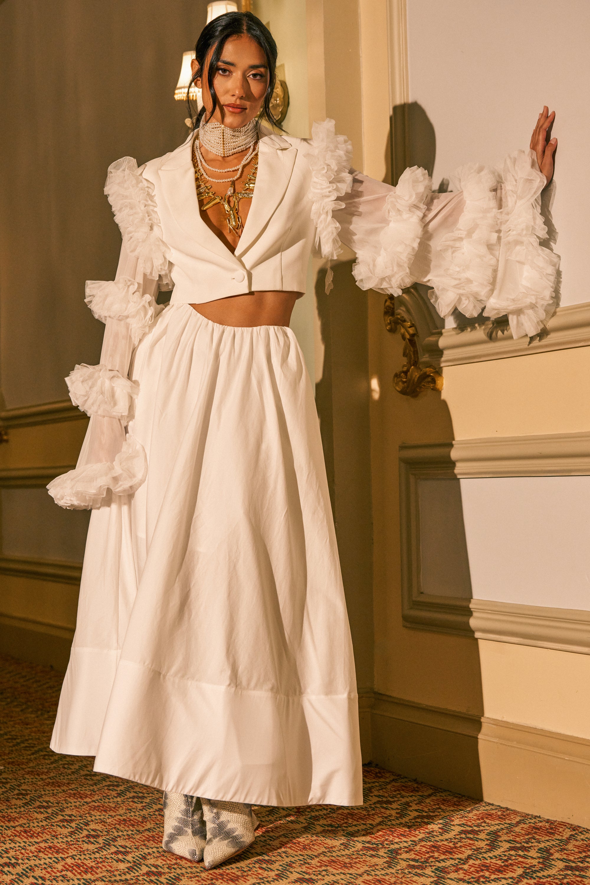 A woman poses indoors wearing the LETS GO PARTY MAXI SKIRT paired with a white crop jacket with ruffled sleeves, layered necklaces, and silver boots. The lighting is warm and elegant.
