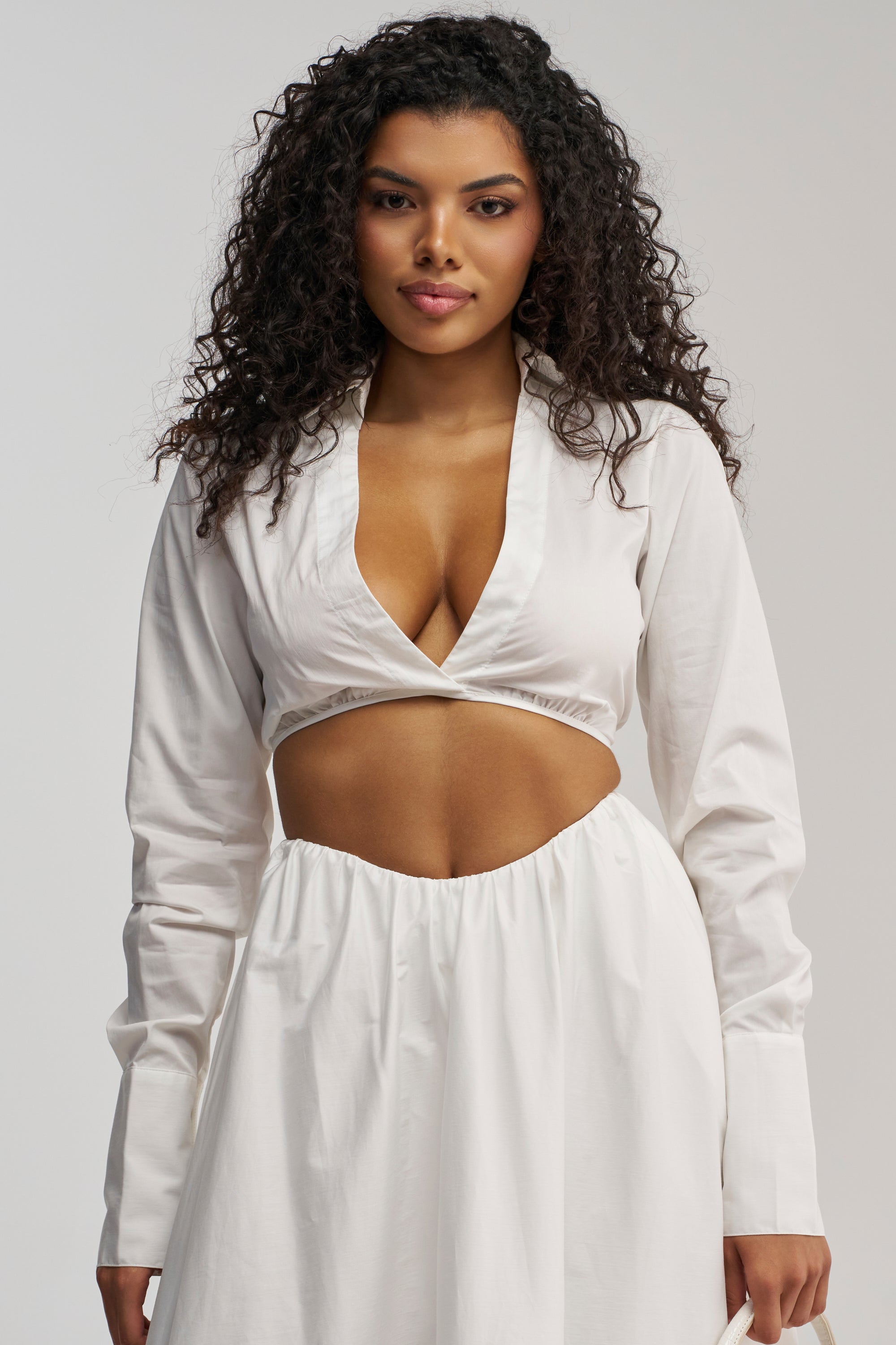 A woman with curly hair stands against a plain background, wearing the LETS GO PARTY BLOUSE—a white, long-sleeve top featuring a plunging neckline and midriff cutout, perfectly reminiscent of a summer date night look.
