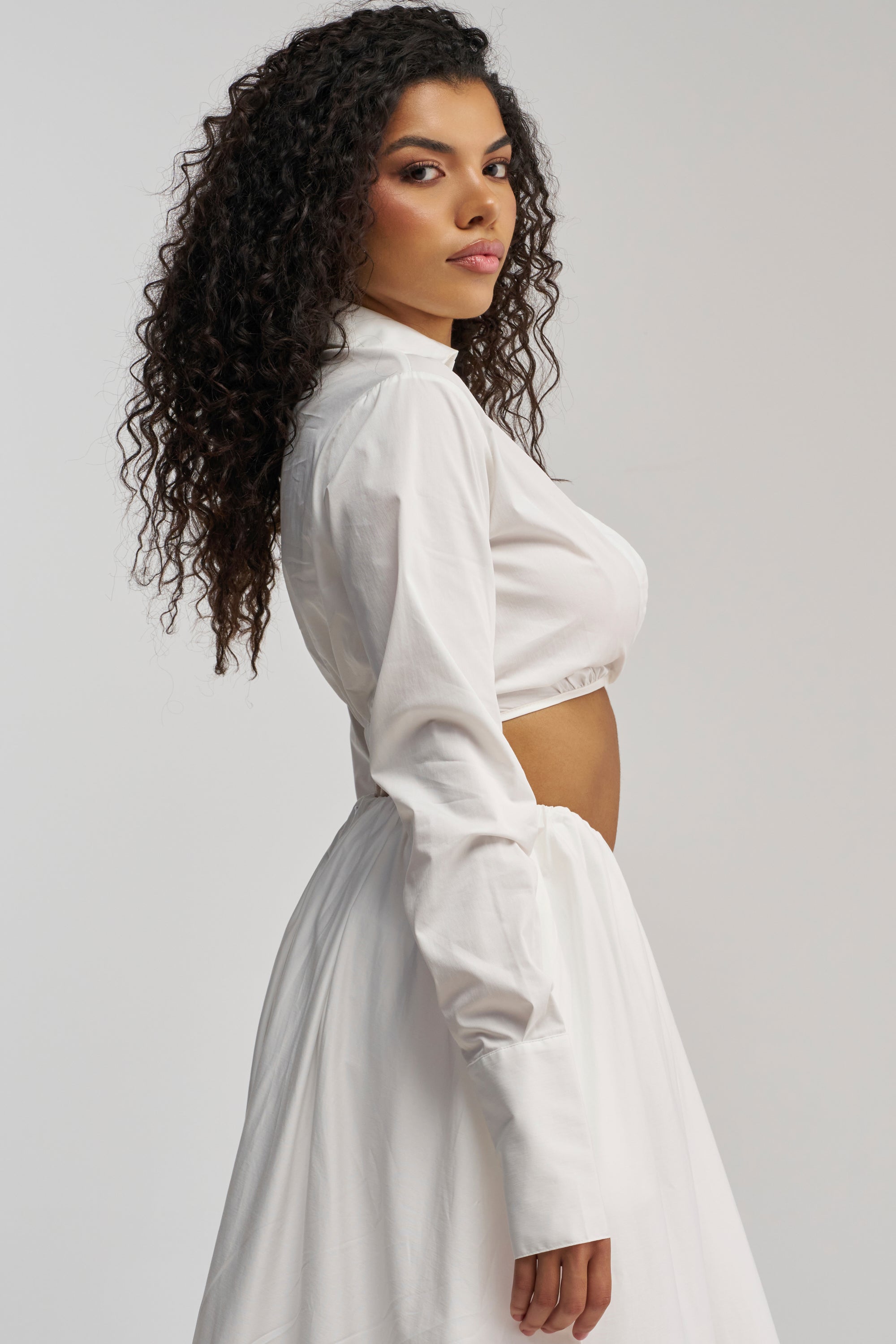 A woman with curly hair poses sideways against a plain light background, wearing the LETS GO PARTY BLOUSE—a white poplin cropped blouse featuring long sleeves and a high neck—paired with a matching skirt.