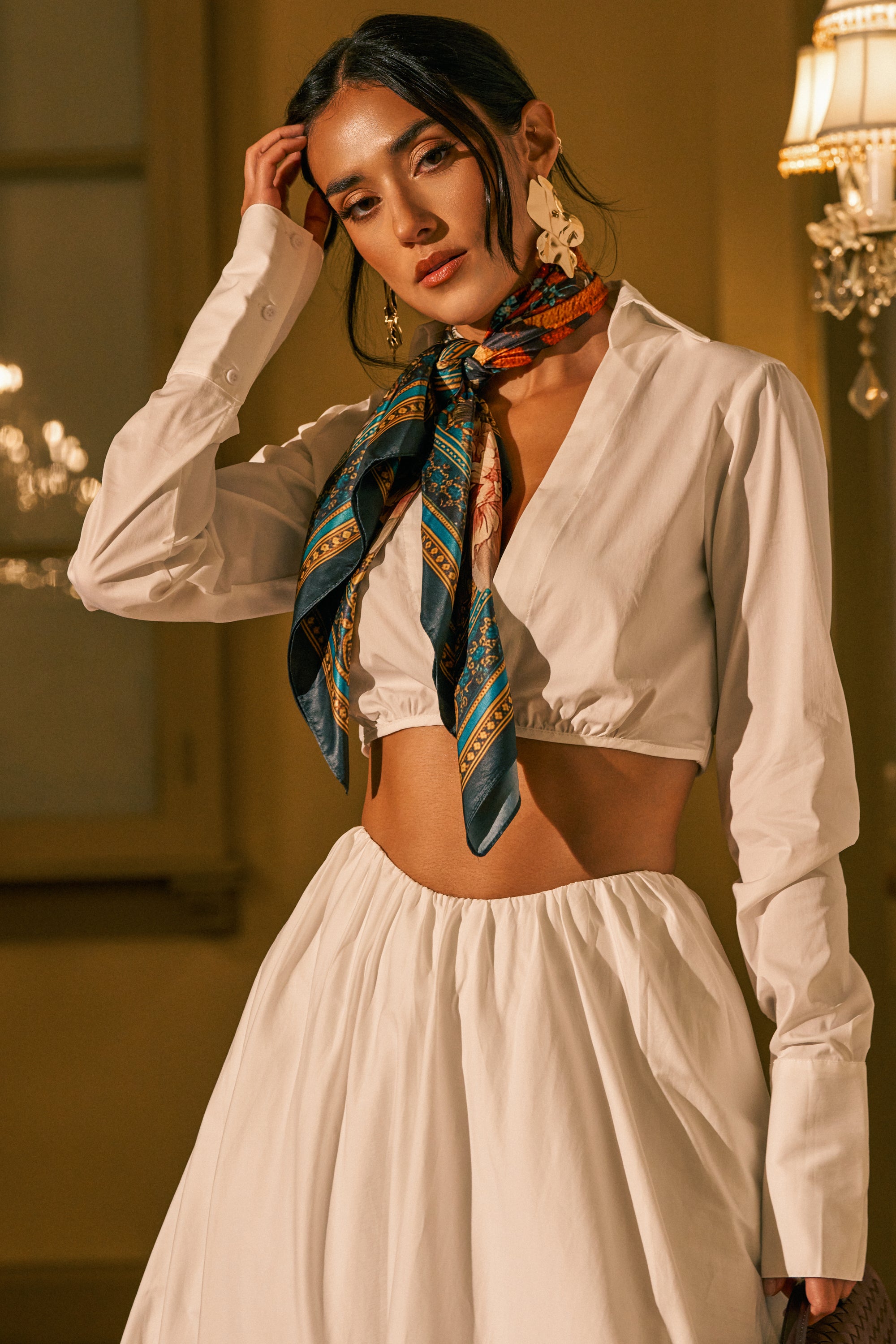 A woman poses indoors in the LETS GO PARTY BLOUSE, a crisp white poplin cropped collared blouse, styled with a matching skirt, patterned scarf around her neck, and statement earrings. She touches her hair and looks confidently at the camera under warm lighting.