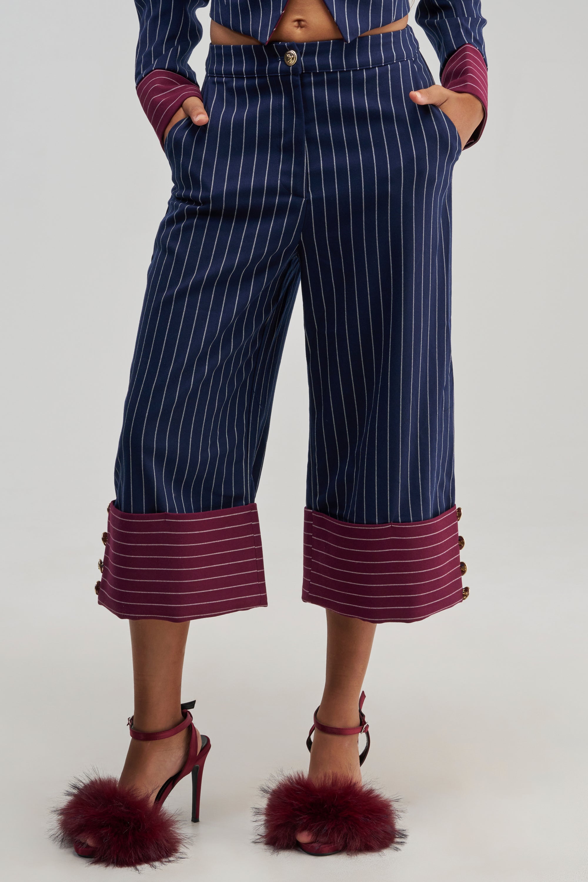 Dressed in the NEVER BETTER PINSTRIPE TROUSER featuring navy pinstripes and bold maroon cuffs, paired with a coordinating blazer detail and maroon high heels with plush accents. With hands casually tucked into pockets and only the lower body in view, this look offers a refined twist on classic office attire.