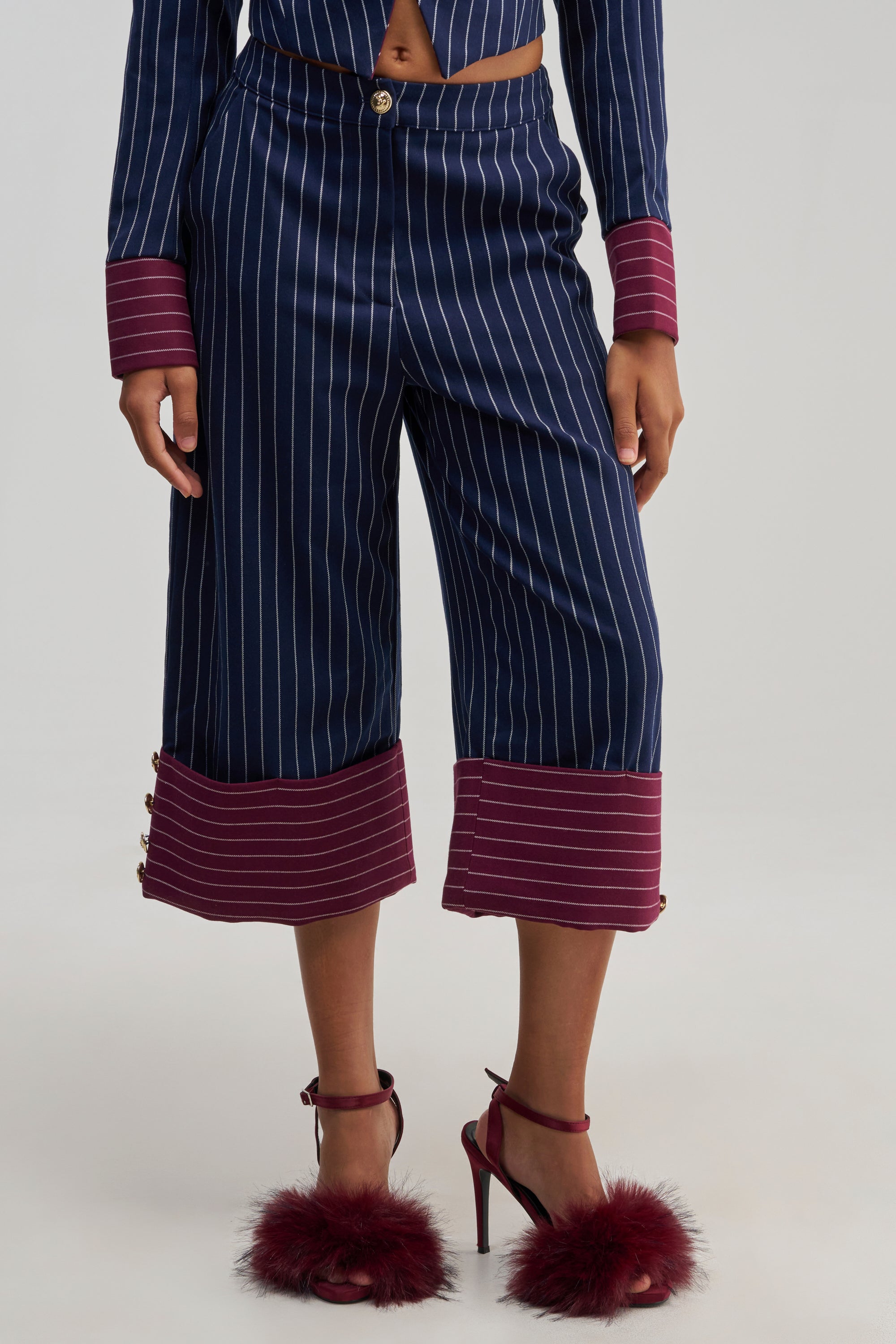 Wearing the NEVER BETTER PINSTRIPE TROUSER, styled as wide-leg navy pinstripe culottes with bold maroon cuffs, paired with a matching top and burgundy high-heeled sandals featuring maroon feather accents.