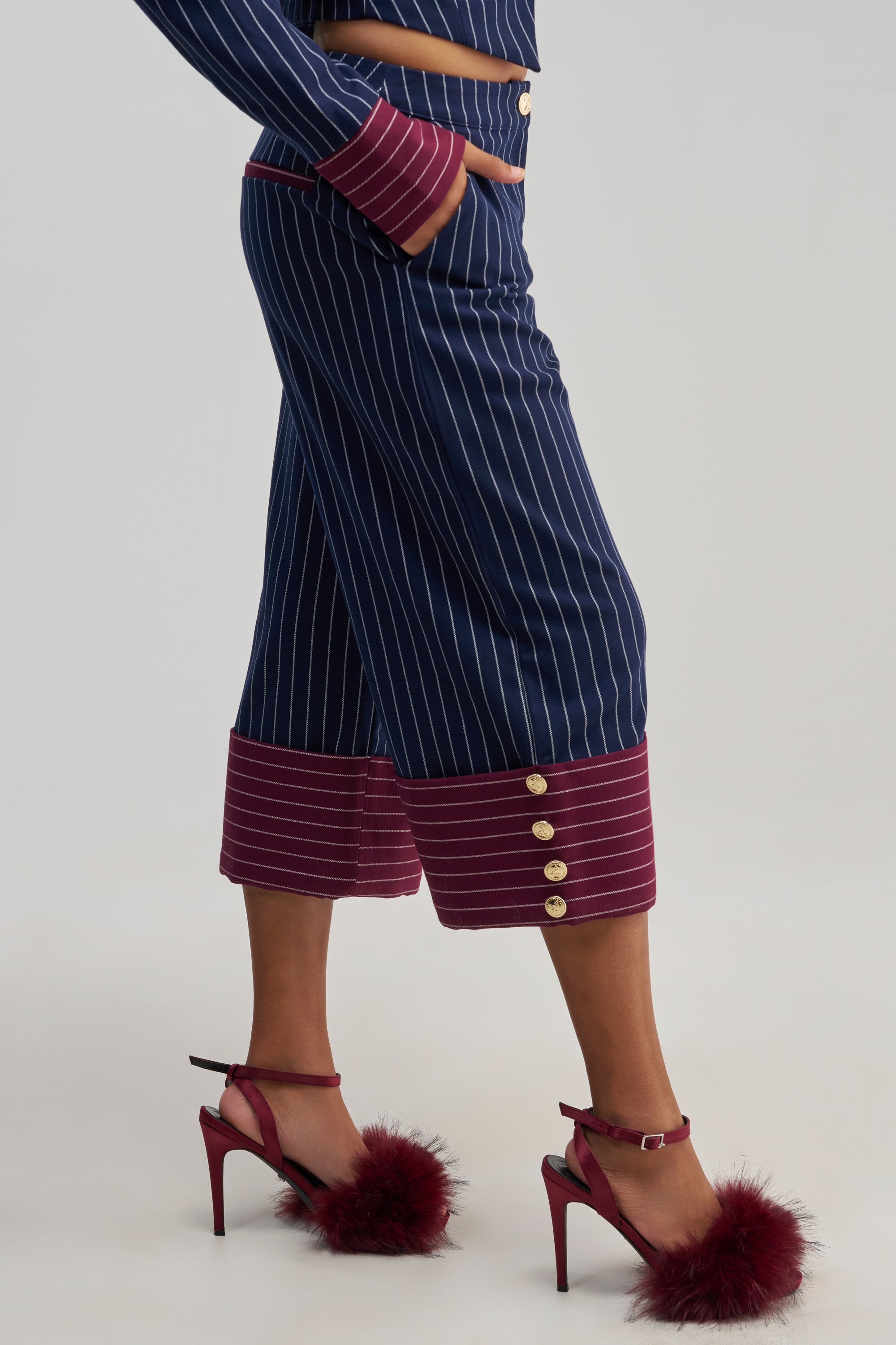 A person is wearing the NEVER BETTER PINSTRIPE TROUSER—navy pinstripe wide-leg culottes with maroon cuffs and gold buttons—paired with a matching top for a sophisticated take on classic office attire. Maroon high-heeled sandals adorned with fluffy maroon accents complete the look, set against a plain light gray background.