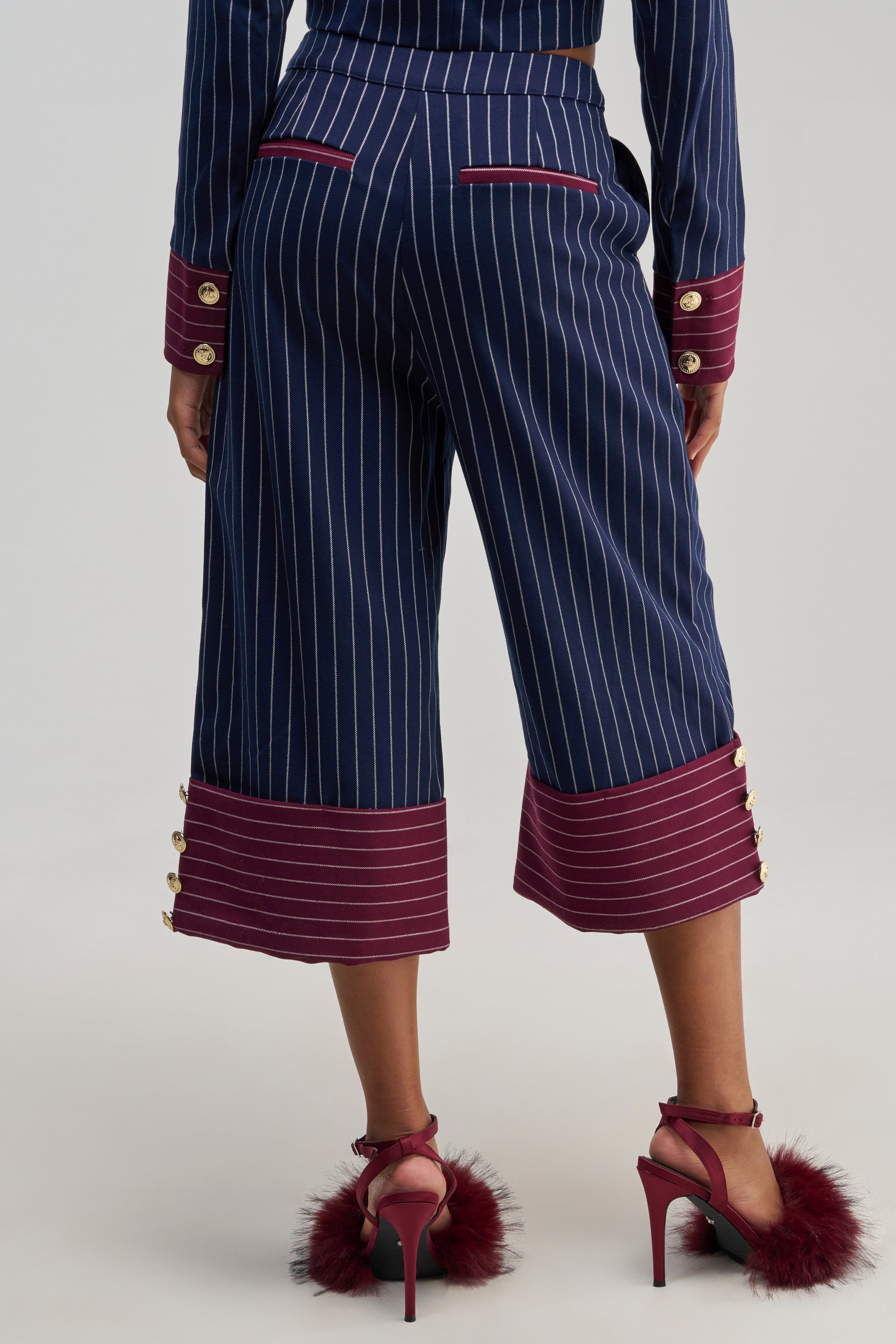 Wearing the NEVER BETTER PINSTRIPE TROUSER—navy cropped pants with maroon cuffs and gold buttons—a person pairs them with matching maroon high heels featuring feather accents. Photographed from behind, the image highlights the chic detailing and bold officewear statement of these modern culottes.