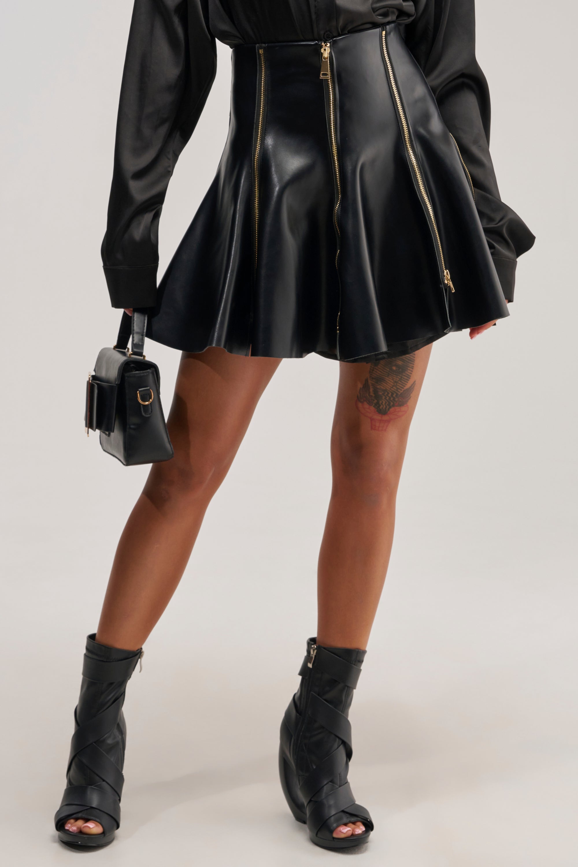 A person wearing the CUTIE PIE ZIPPER MINI SKIRT in black faux leather with zippers, paired with a black long-sleeve top, heeled black sandals, and holding a small black bag. A tattoo is visible on their thigh.