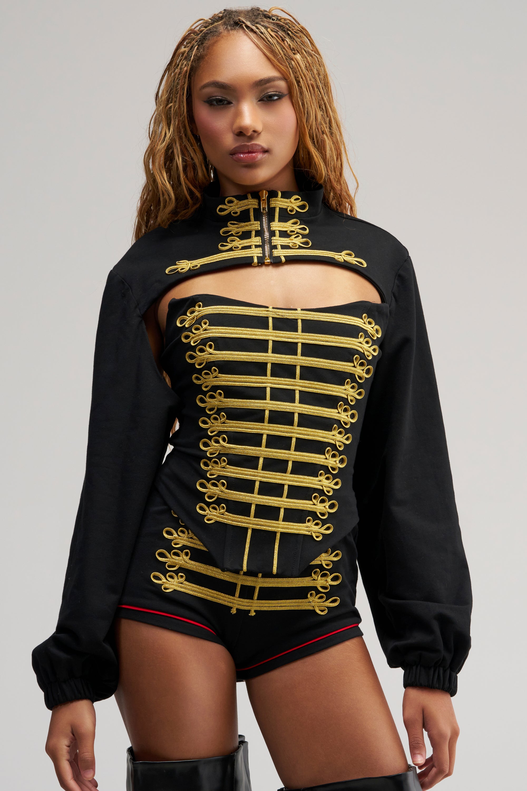A person wears a black high-neck outfit featuring gold decorative trim, long sleeves, and a chest cutout, layered with the HAUTE COMMAND BOLERO—a sleek black military-inspired bolero. The ensemble is completed with black boots and set against a plain, neutral background.