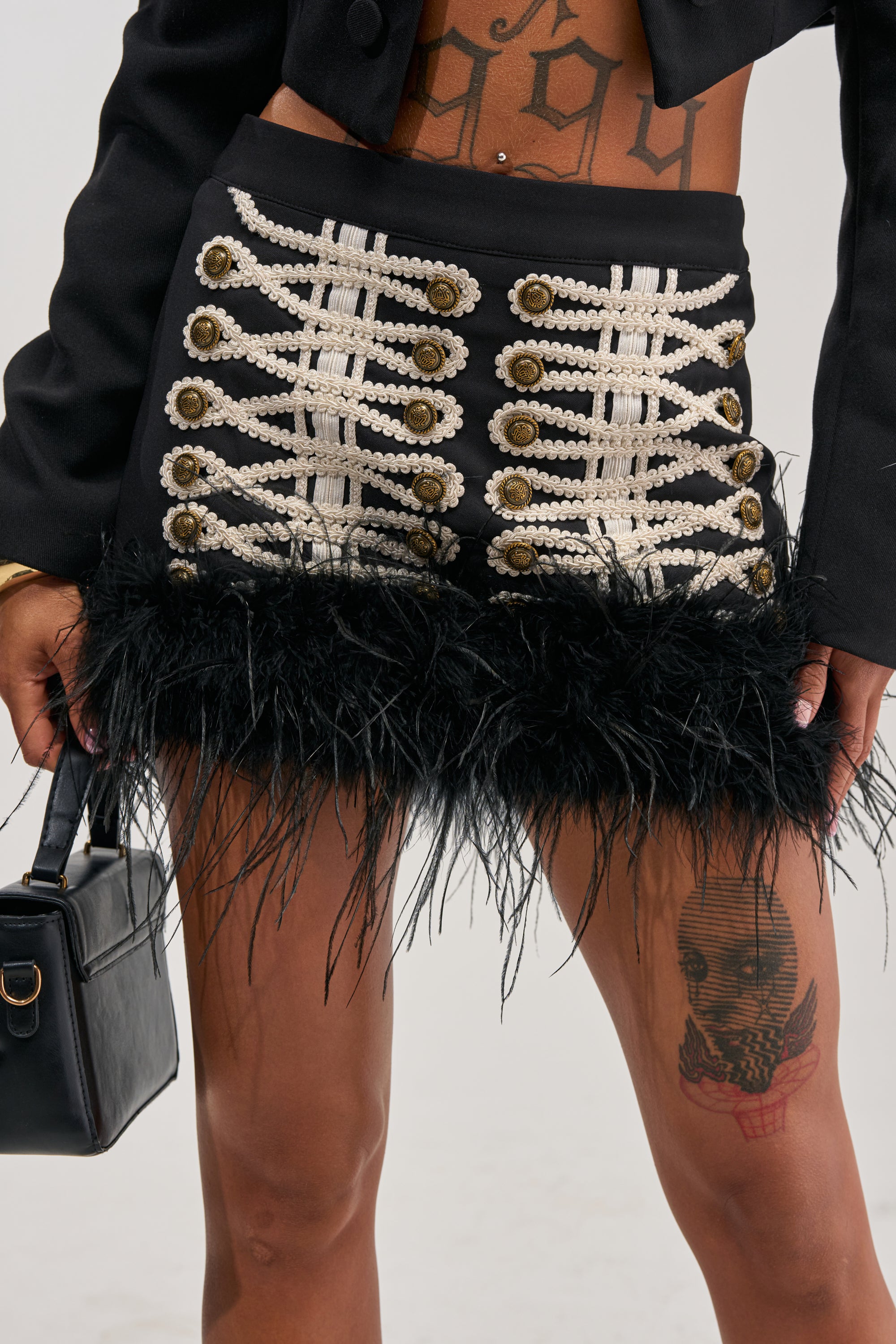 A close-up shot shows a person wearing the GET THE VIBE MINI SKIRT, distinguished by its military-inspired design with gold buttons, white braided trim, and a statement feather-trim hem. The look reveals a thigh tattoo and a visible midriff, while the person accessorizes with a black handbag for an elevated utility-chic vibe.