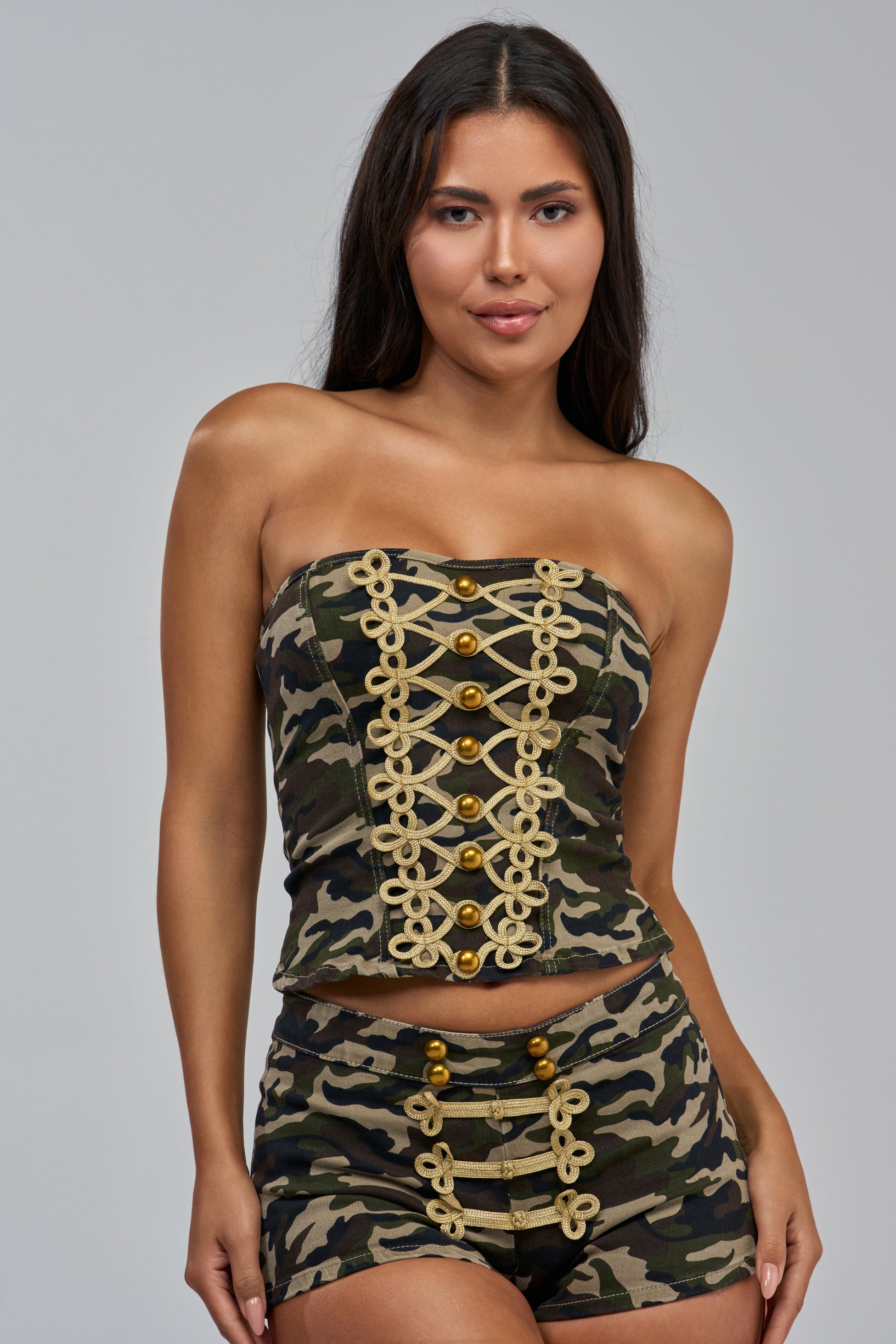 A woman stands against a plain gray background, smiling slightly as she models the DRILL ME DOWN CORSET—a camo military strapless corset-style top with gold decorative buttons and trim—paired with matching shorts.
