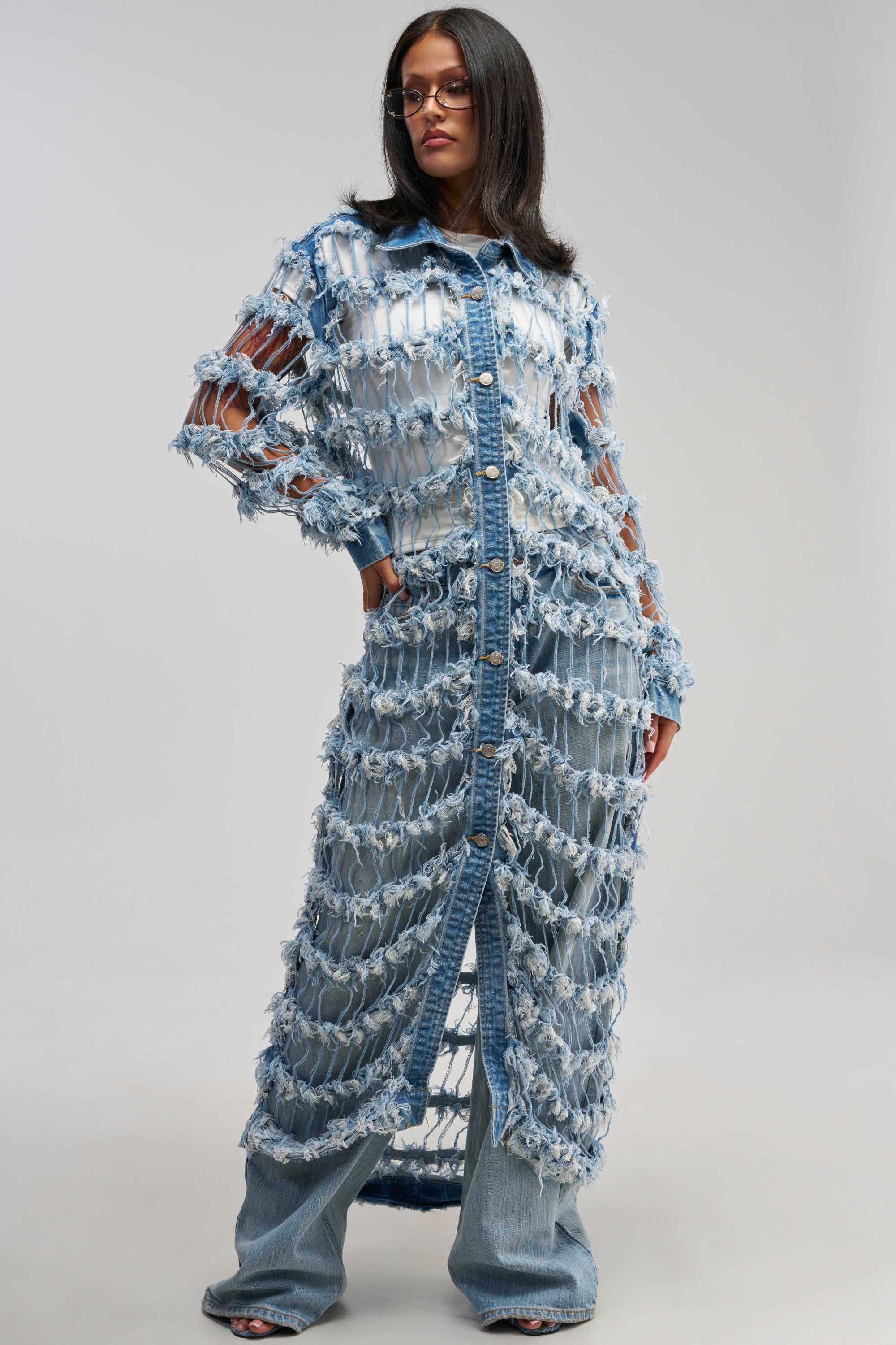 Wearing the GOOD TIME NOT A LONG TIME DENIM TRENCH—a long, avant-garde distressed denim coat with large cutouts—layered over a white shirt and denim pants, this individual stands confidently against a plain backdrop, making a bold streetwear statement.