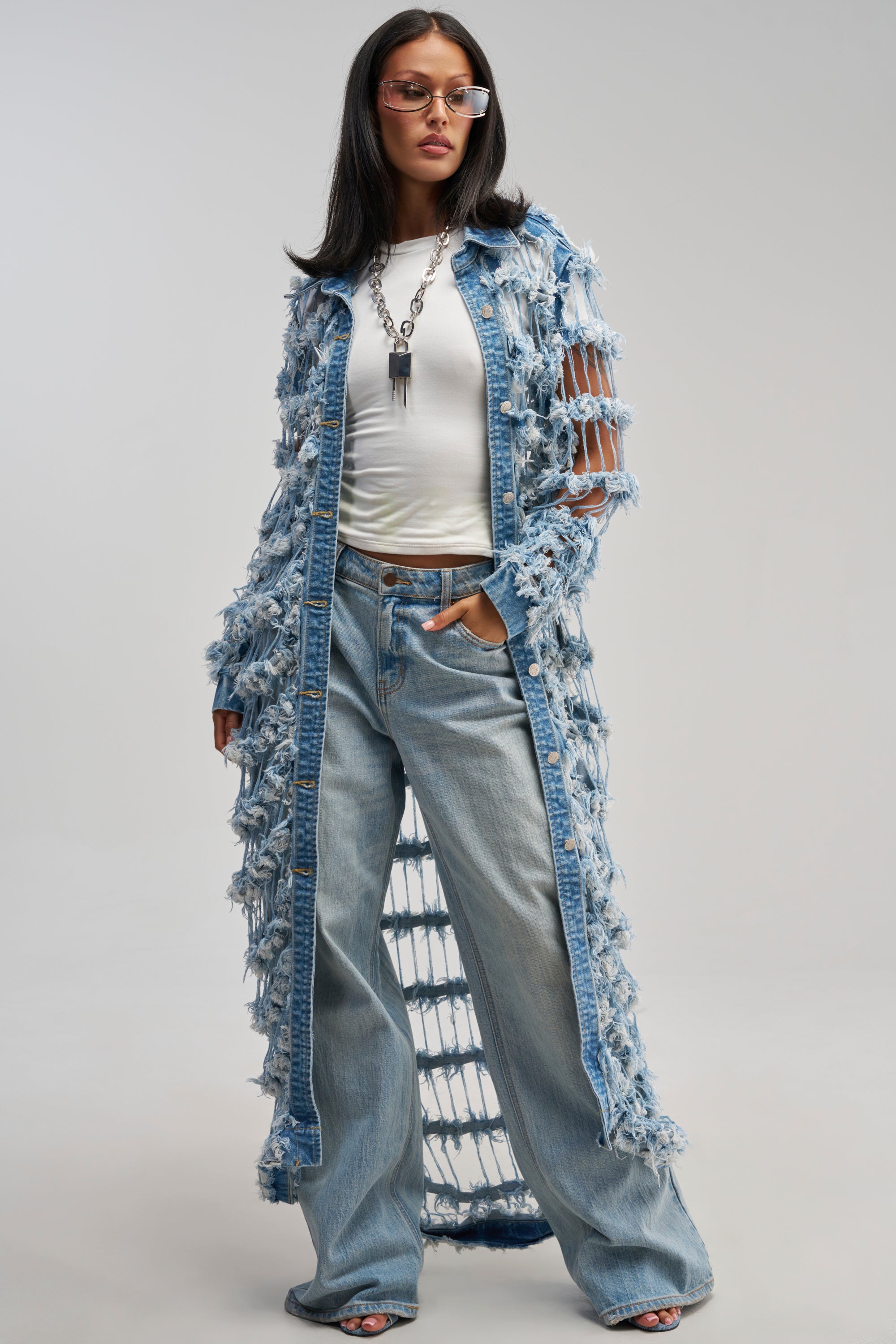 A woman wearing glasses, a white t-shirt, light blue wide-leg jeans, and the GOOD TIME NOT A LONG TIME DENIM TRENCH with avant-garde distressed details and fringe poses confidently against a plain backdrop, effortlessly blending streetwear with contemporary style.