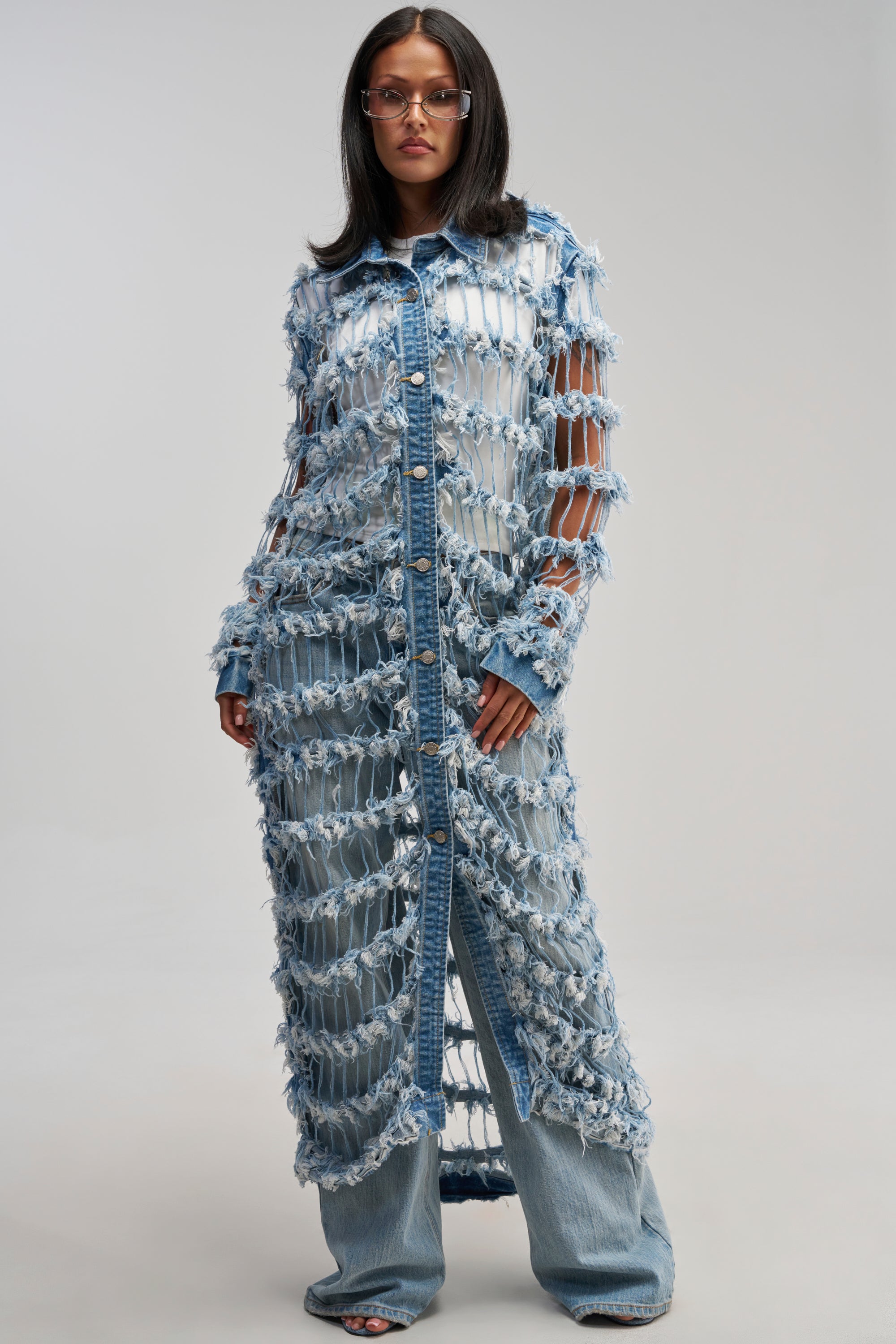 A woman wears the GOOD TIME NOT A LONG TIME DENIM TRENCH, an avant-garde coat designed with a distressed finish, frayed edges, and bold cut-out gaps. Styled over a white top and wide-leg jeans, the trench features front buttons that amplify its streetwear vibe. She completes her look with glasses and stands against a plain background.