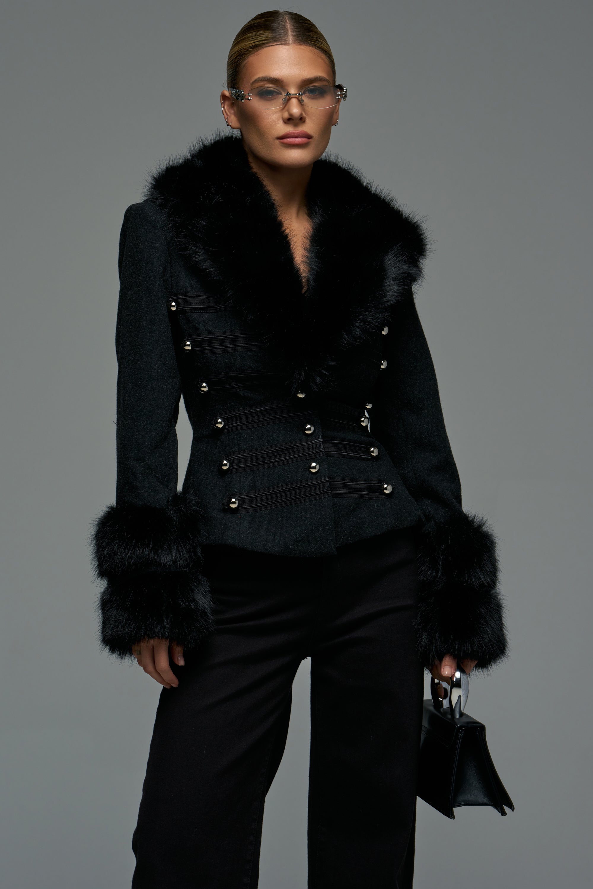 A woman wears THE VISION FUR TRIM BLAZER, a black faux fur blazer with a fur collar, fur-trimmed cuffs, and military-inspired silver button detailing.