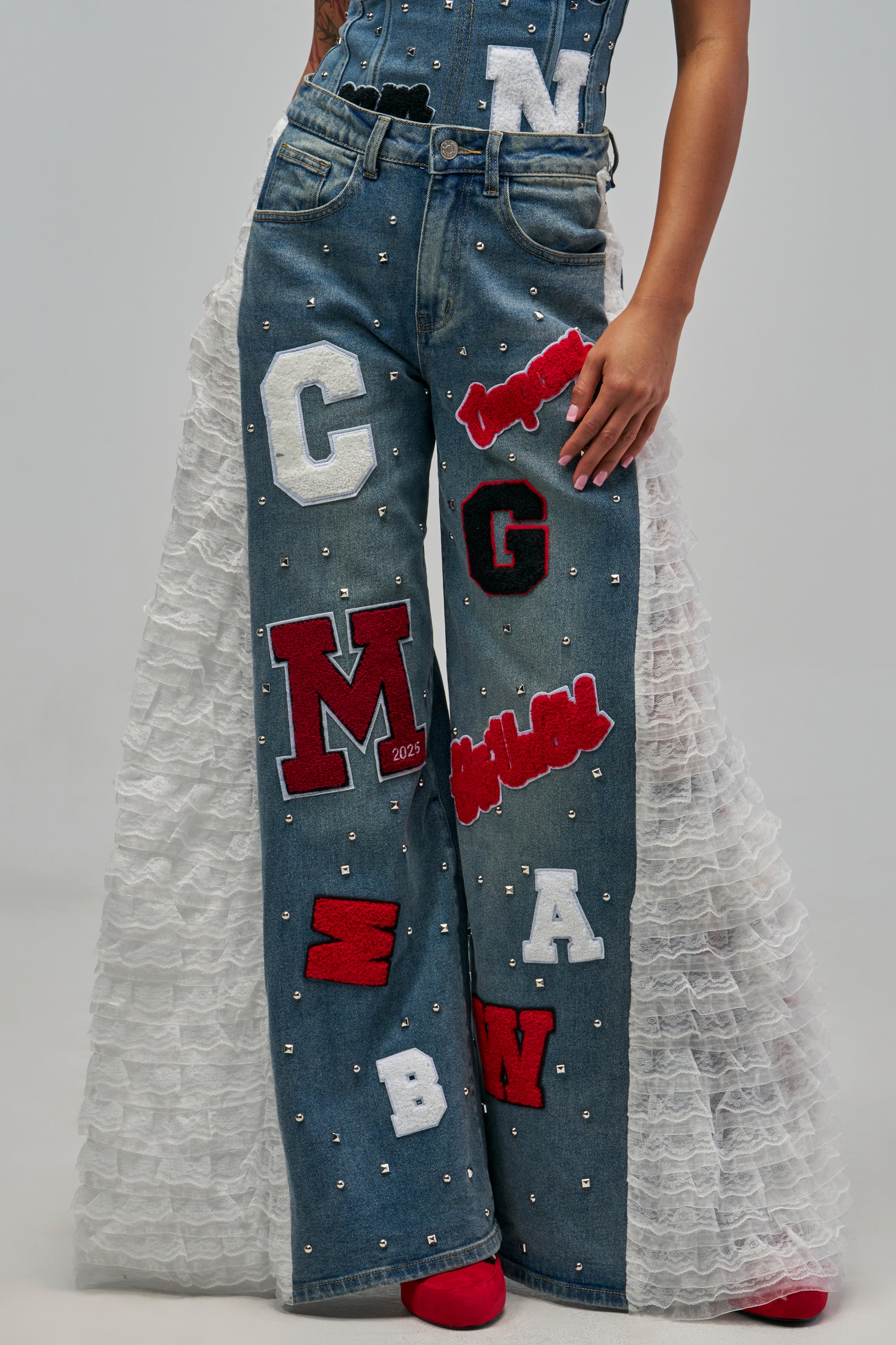 GAME DAY GIRLY DENIM medium blue wide-leg jeans featuring metal studs, varsity-inspired letter patches, and long white lace ruffles along the side seams.