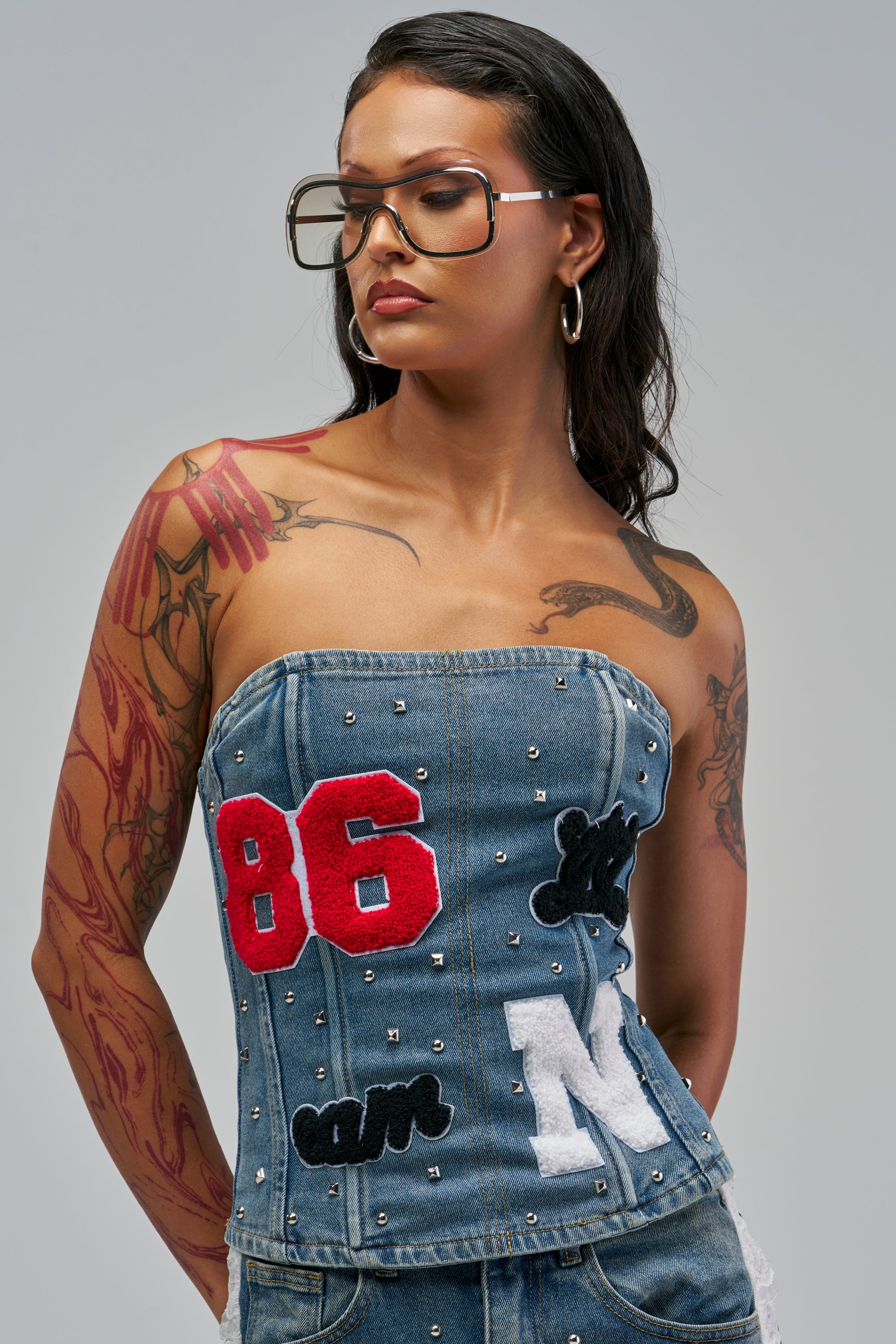 The GAME DAY GIRLY CORSET is a sporty, varsity-inspired denim top decorated with assorted fabric patches and small metal studs.
