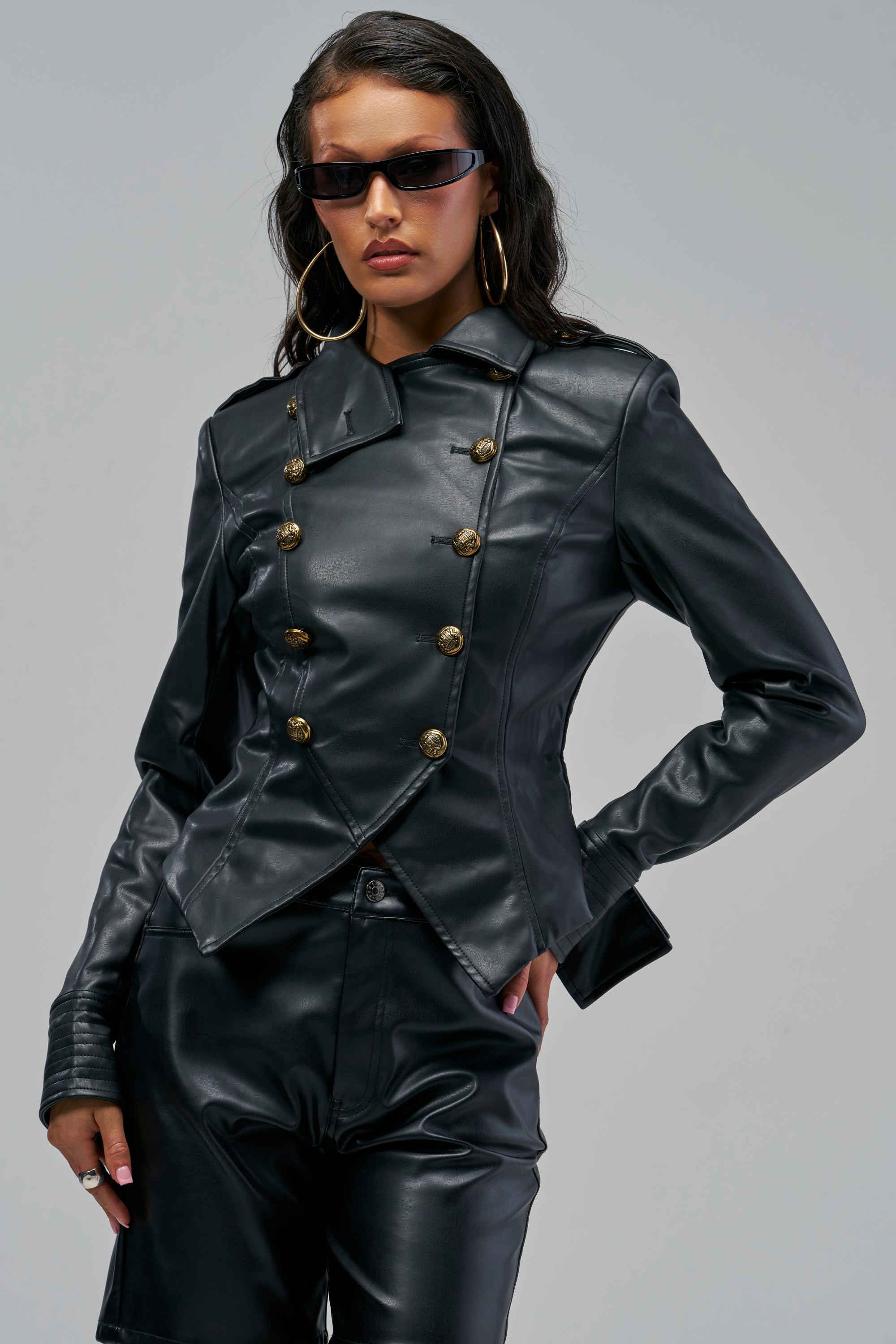 A woman wears a black NAYA FAUX LEATHER BLAZER with gold buttons and matching trousers, creating a sharp statement look.