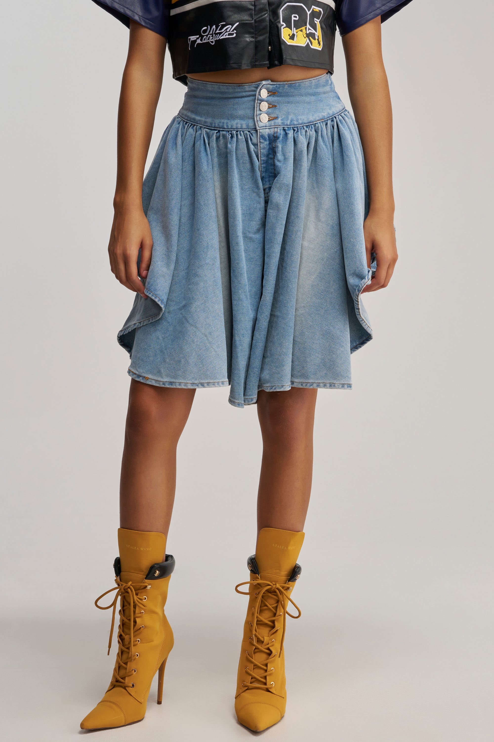 Wearing the COOLEST GAUCHO PANT—a blue, high-waisted, button-front denim piece with draped sides reminiscent of light wash gauchos—paired with brown lace-up high heel boots and a cropped black jacket, this look brings an elevated streetwear vibe. The image is cropped at the shoulders.