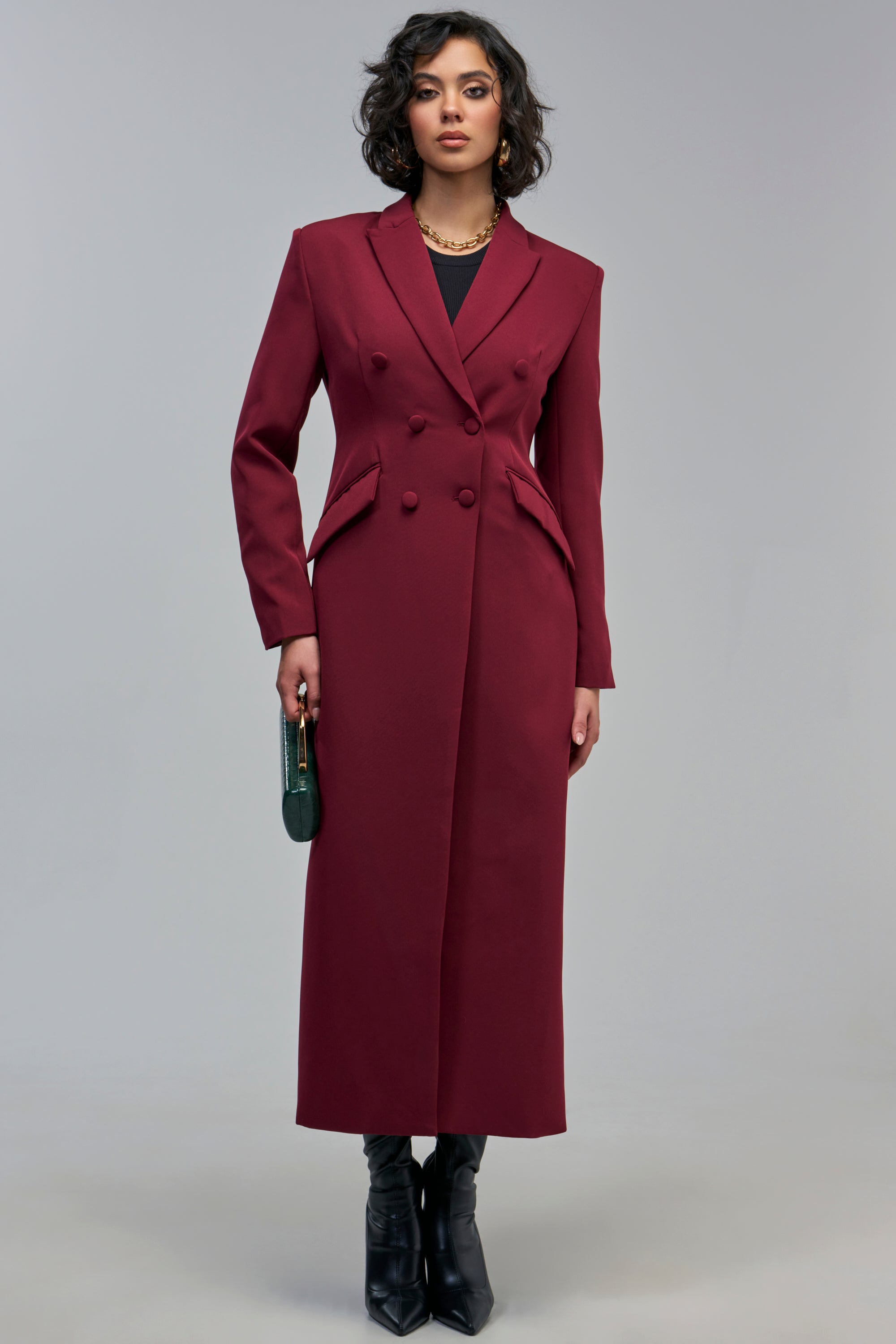 A person wears a long, double-breasted, dark red NO QUESTIONS ASKED TRENCH over a black shirt and black boots, holding a small green handbag.